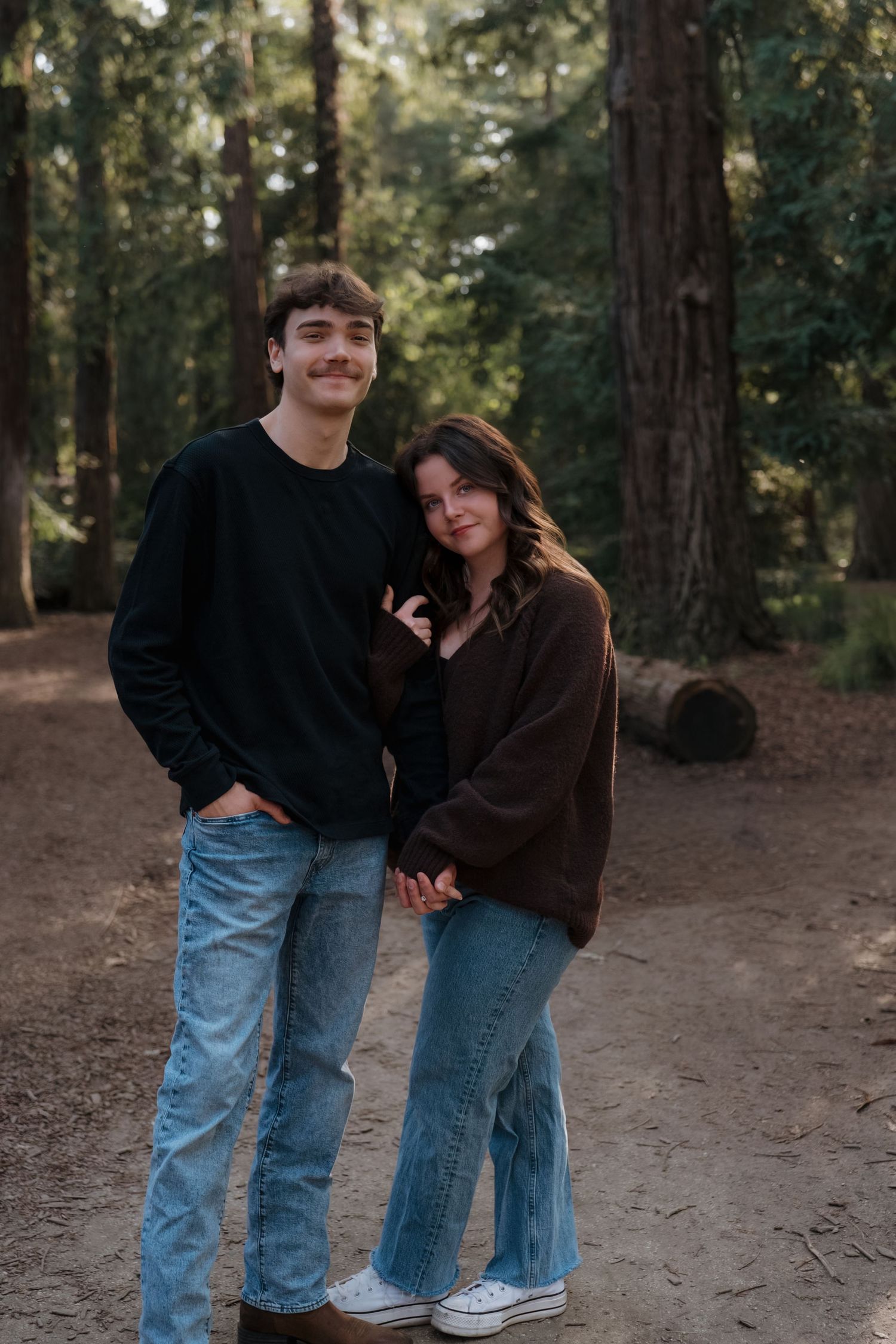 Young couple posing together on a forest trail surrounded by tall redwood trees, wearing casual jeans and sweaters.