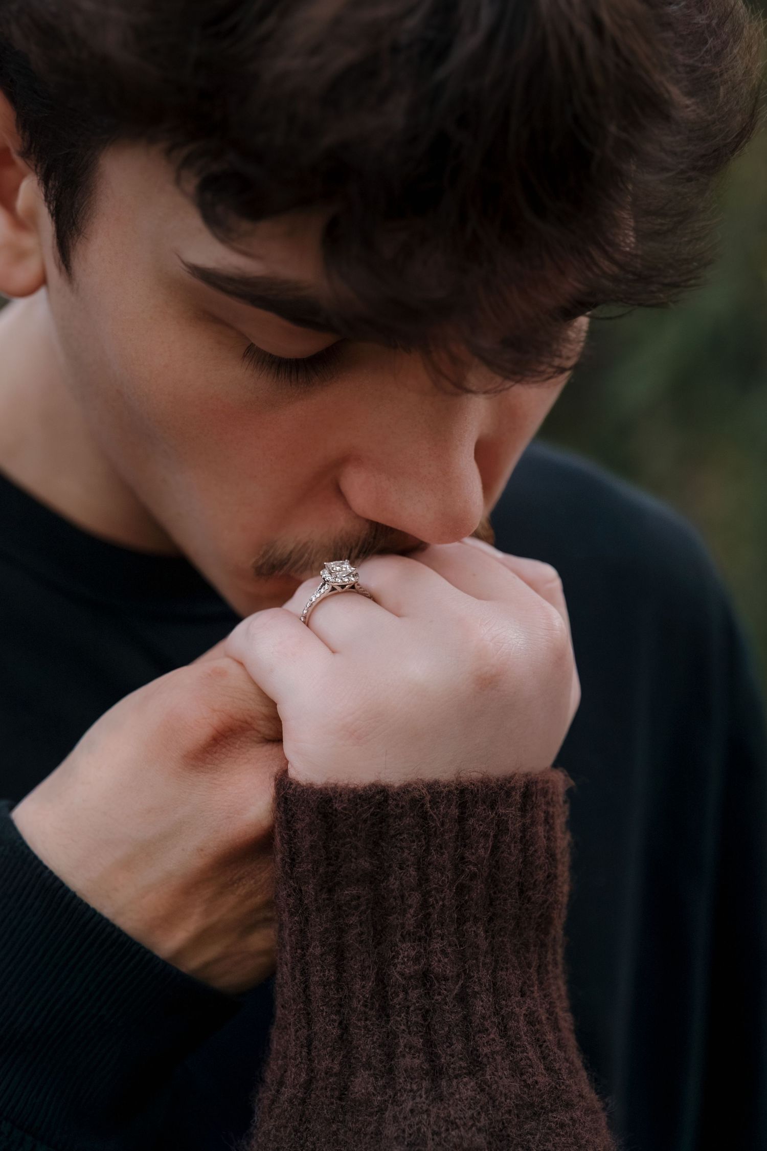 Young man with dark curly hair kissing a hand wearing a diamond engagement ring, romantic proposal moment.