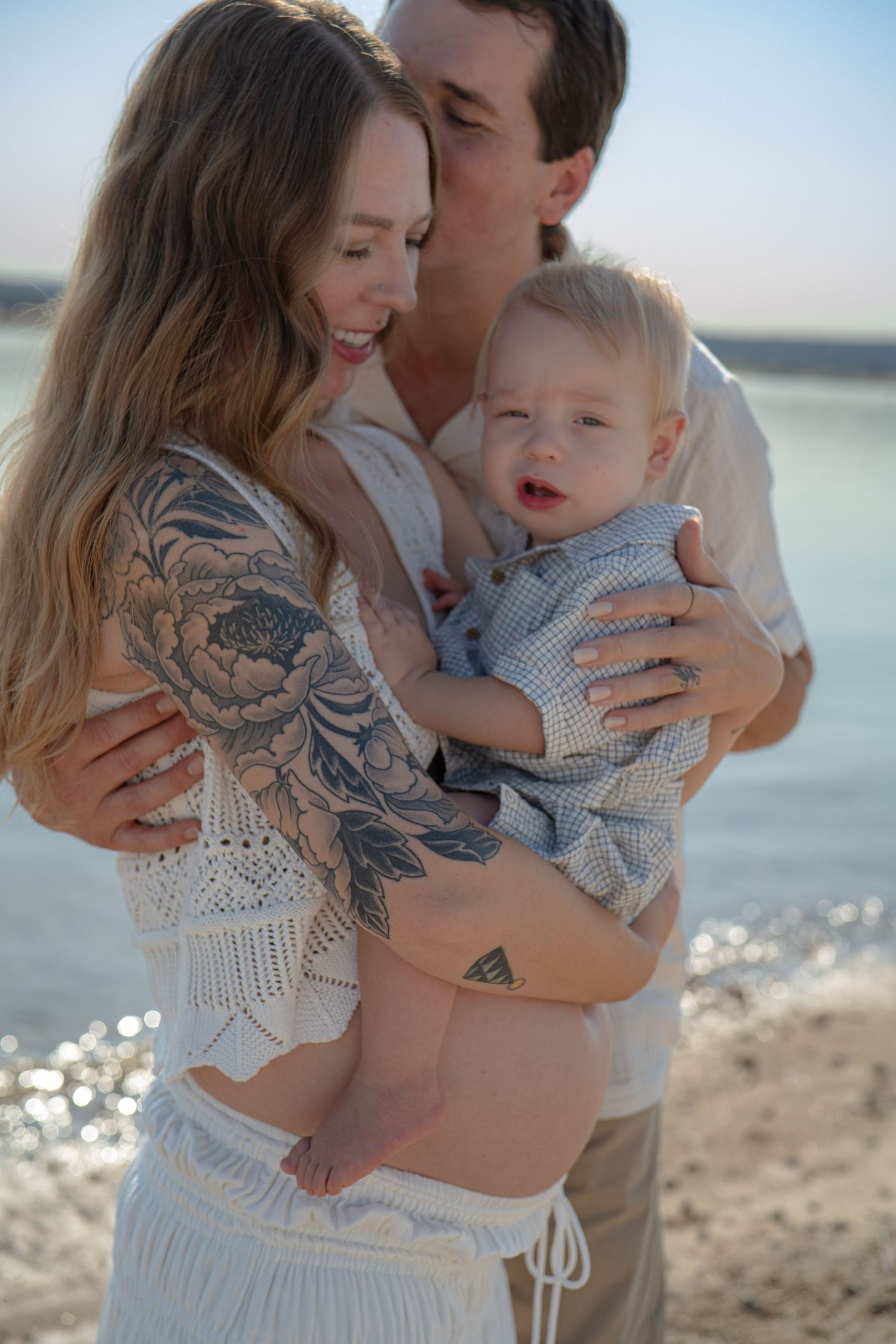 Family portrait at beach with baby wearing blue outfit and parent showing tattoo.