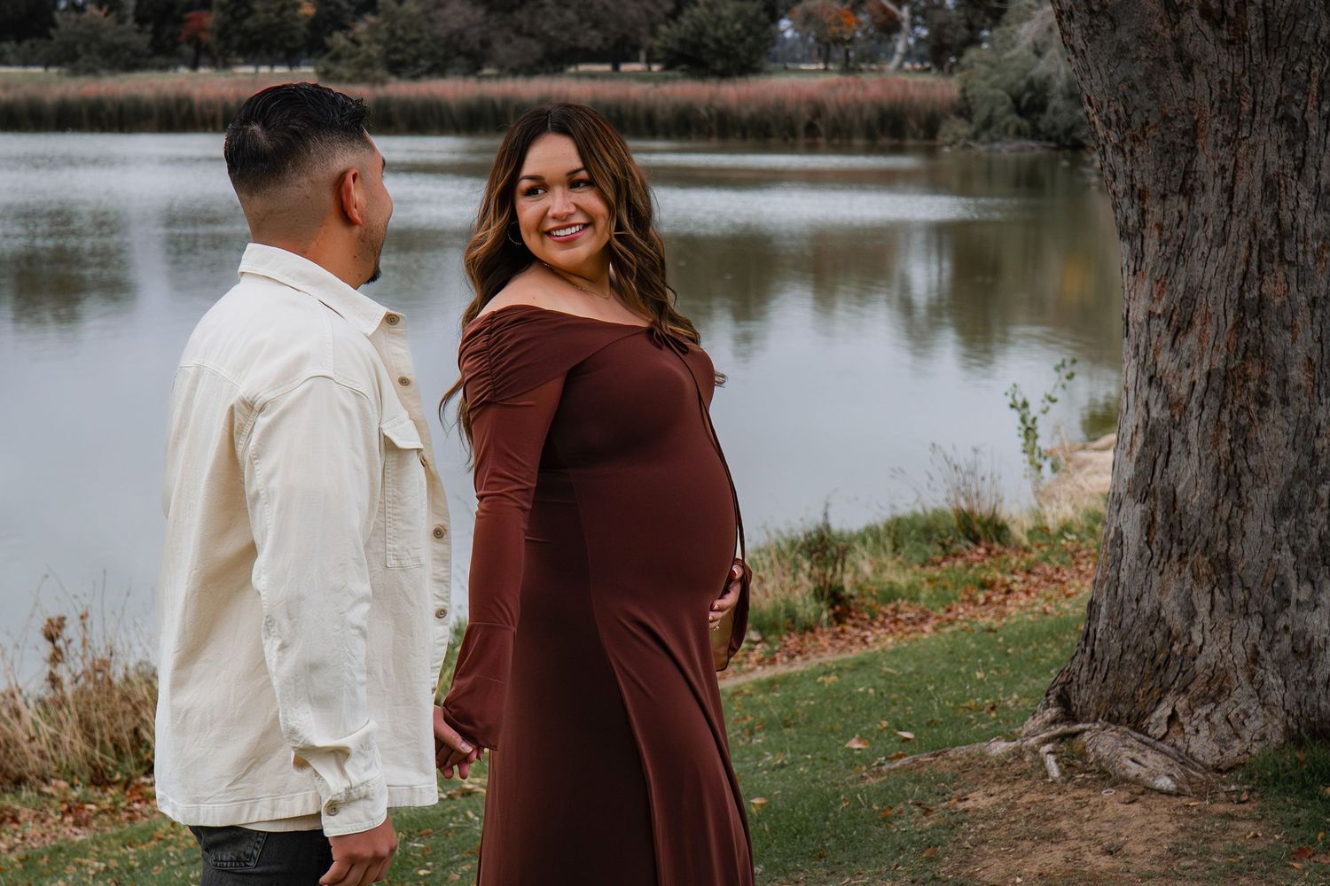 Couple poses by a serene lake surrounded by autumn trees at sunset.
