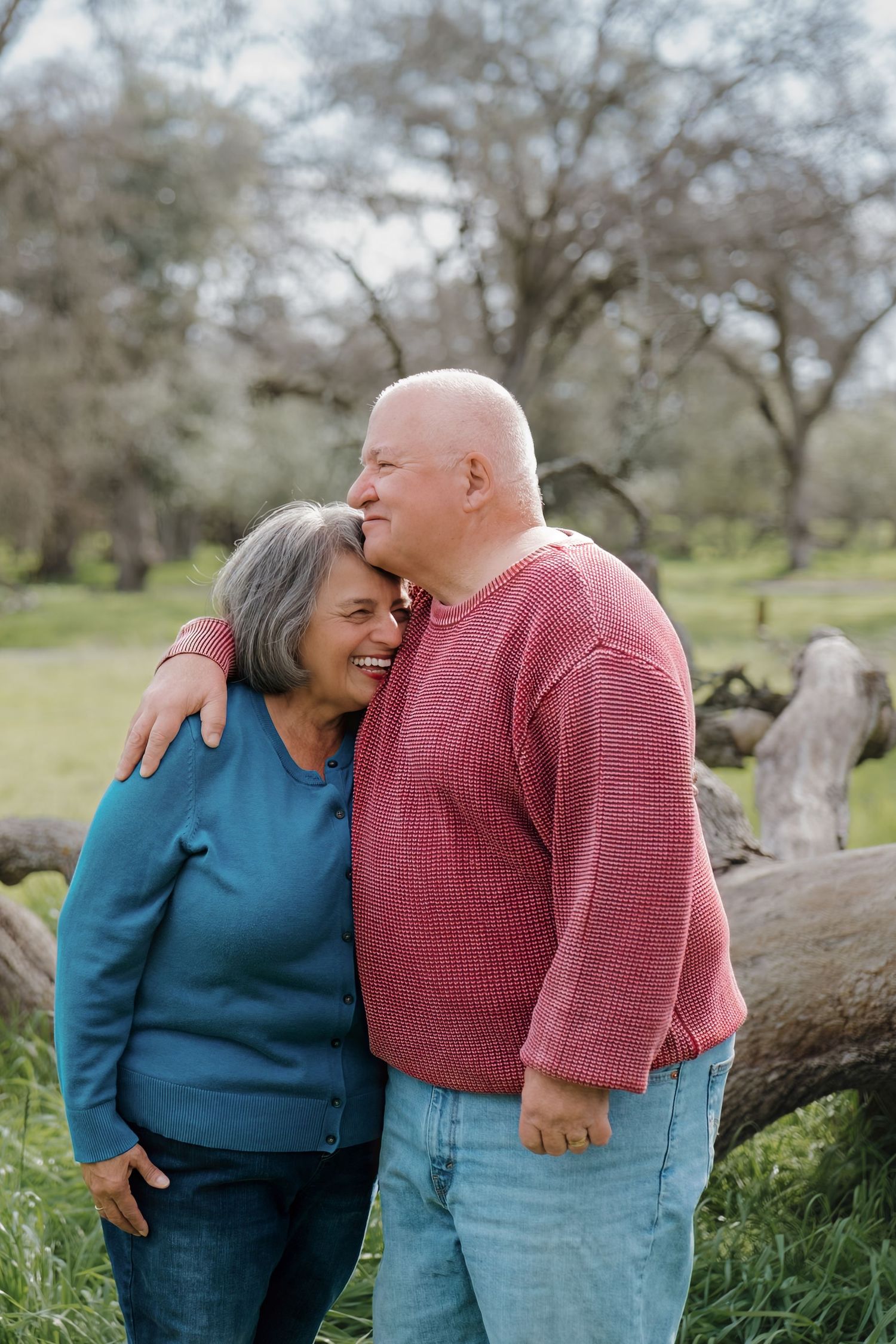 A couple in blue and pink sweaters share intimate moments while sitting on fallen logs in a natural outdoor setting.