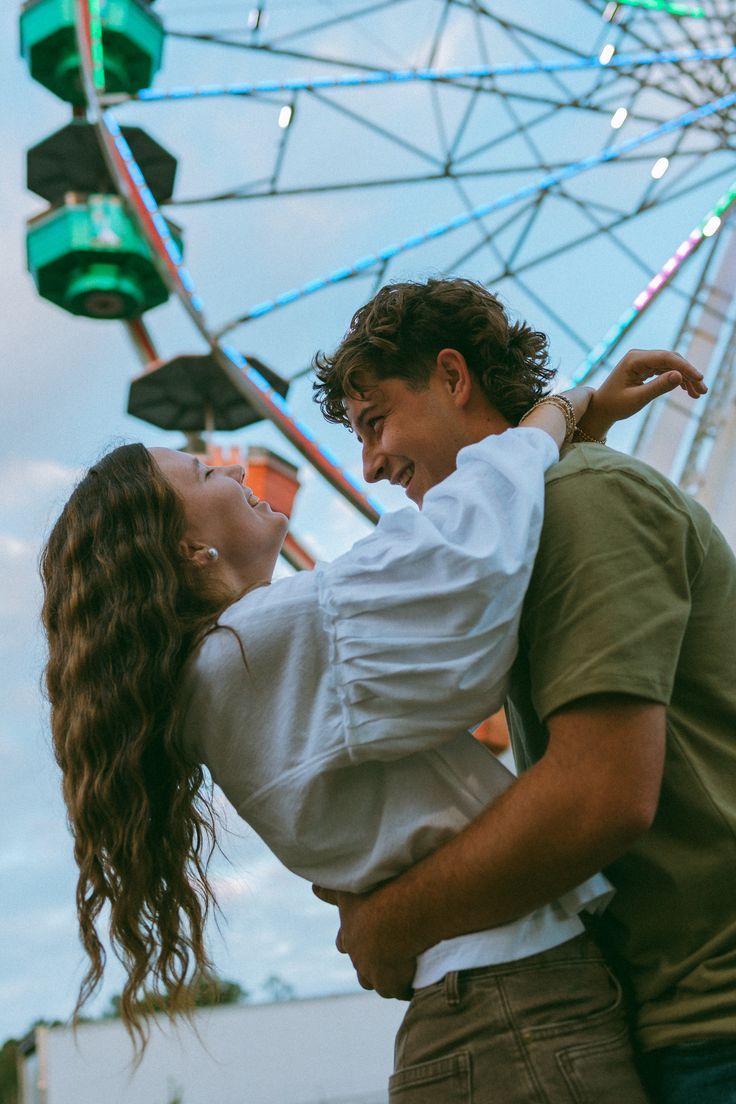 A joyful couple embracing and laughing together in front of a colorful Ferris wheel at an amusement park.