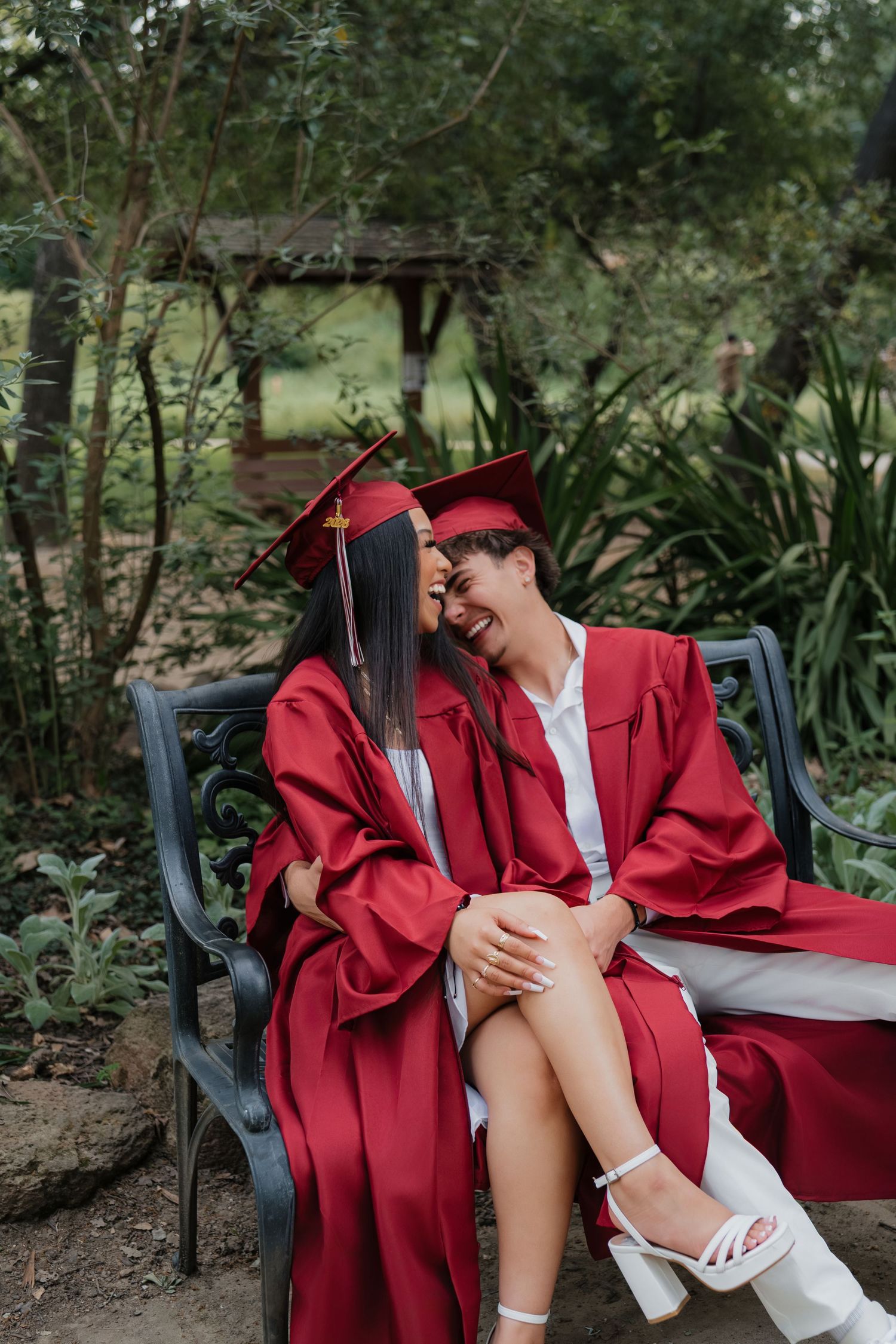 Two graduates in red caps and gowns sit on a bench laughing together in a lush garden setting.