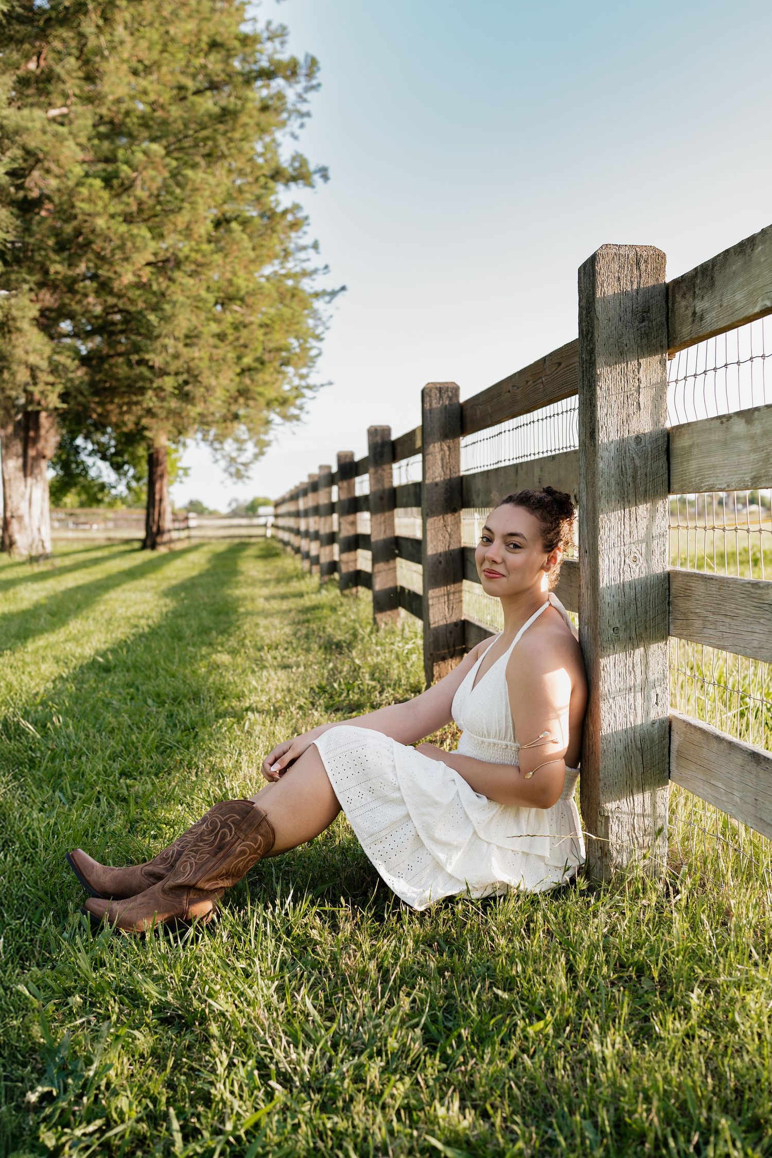 Young woman in white dress and cowboy boots sitting against a wooden fence on green grass at a sunny ranch.
