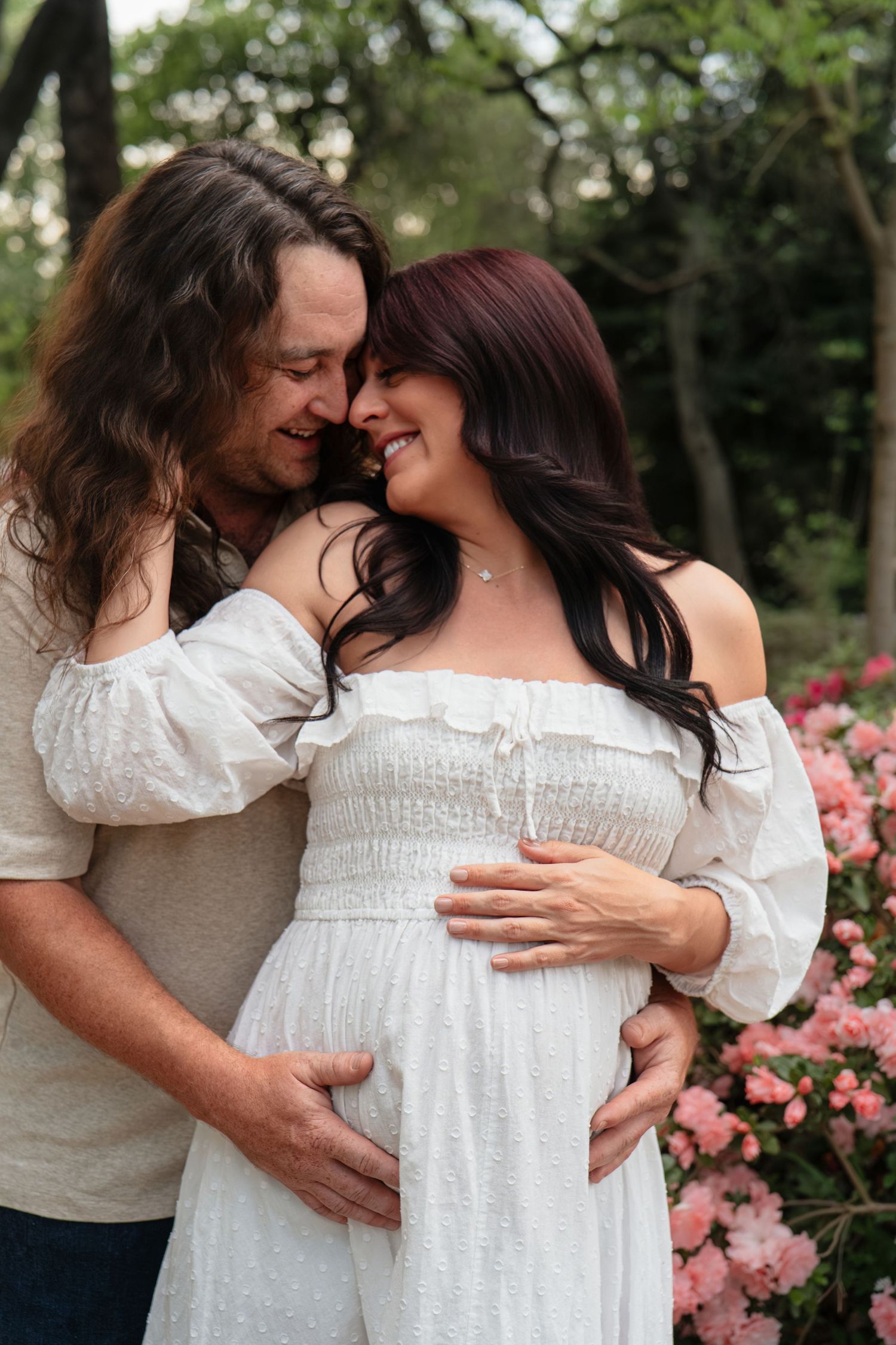 Couple sharing tender moment outdoors, pregnant woman in white off-shoulder dress with partner embracing her baby bump near pink flowers.