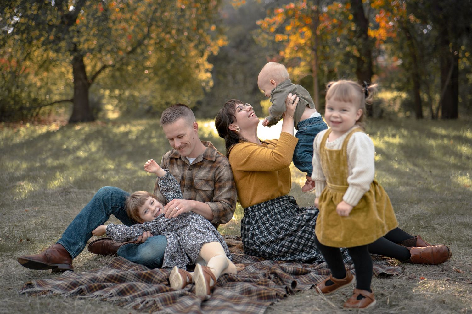 A family group lies on a brown blanket outdoors during an autumn photoshoot in a wooded setting.