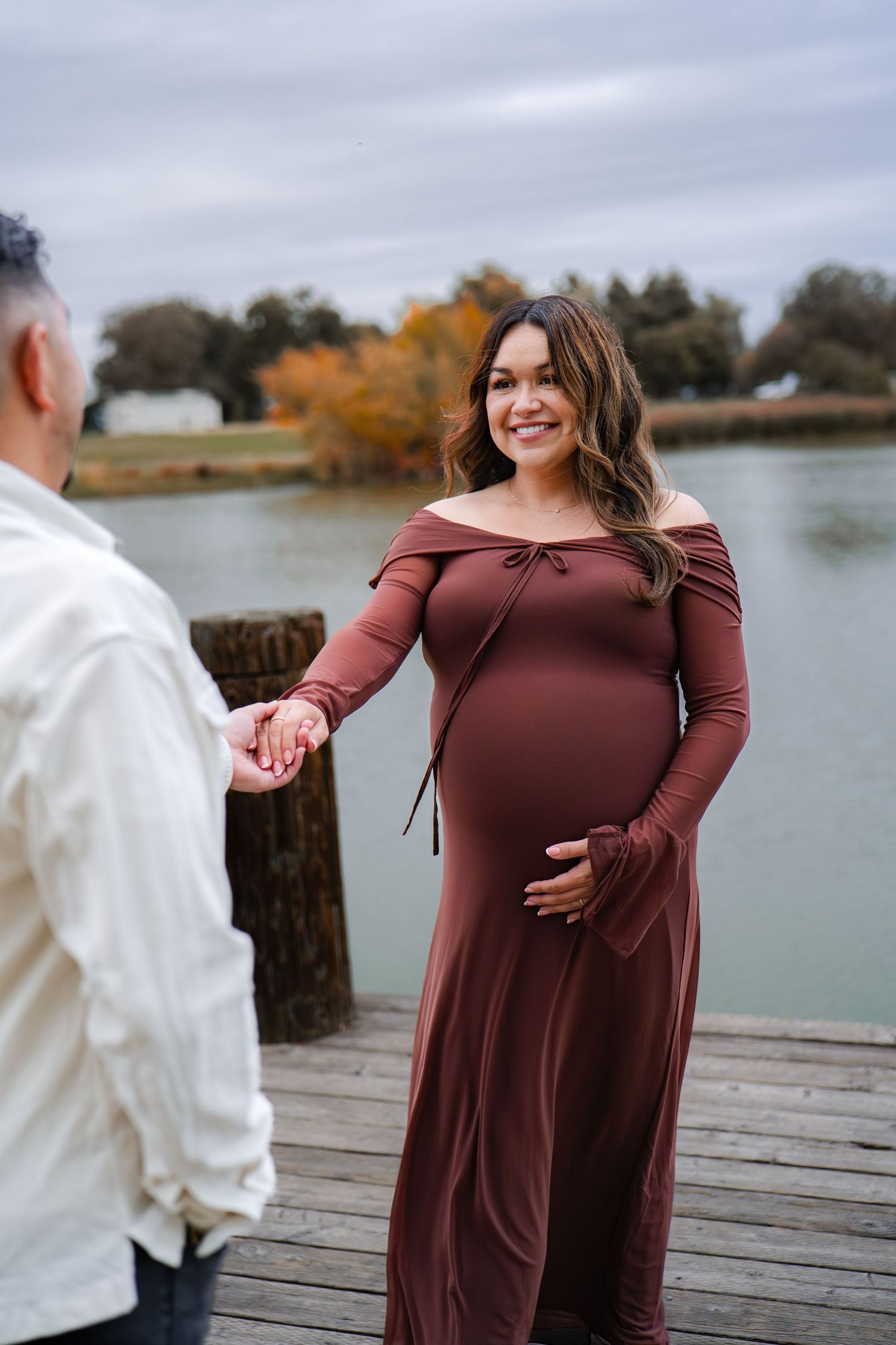 Maternity photos on wooden dock showing woman in burgundy dress by autumn lake in Roseville, CA