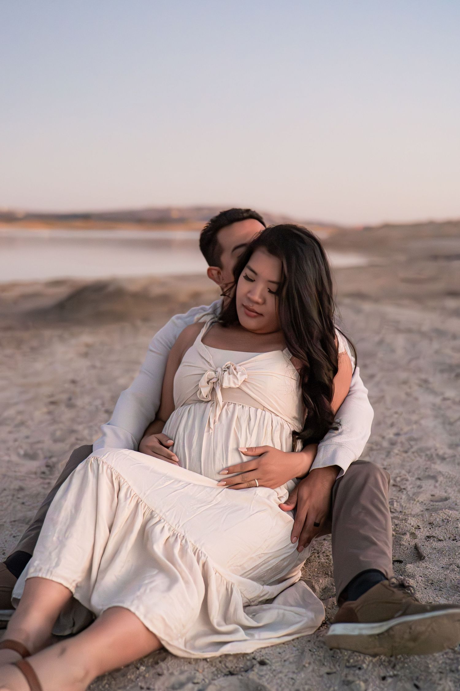Couple embracing on beach at sunset with pregnant woman in white dress.