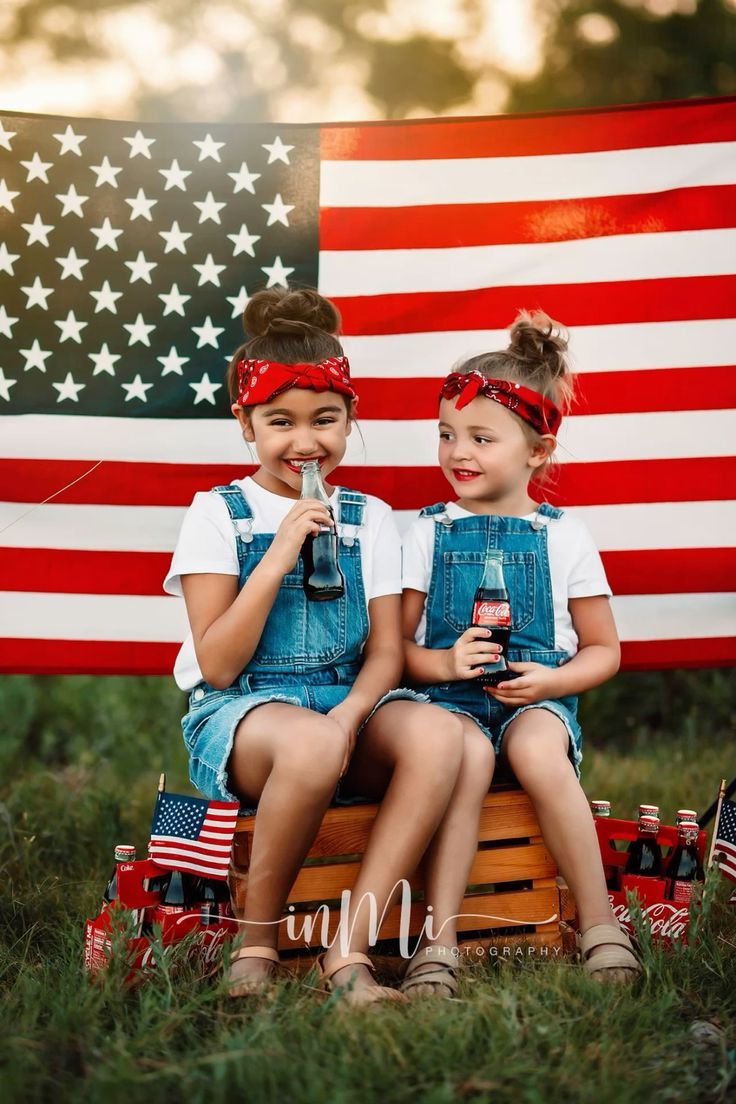 Two girls in denim overalls and red bandanas sit on a crate with Coca-Cola bottles before an American flag.