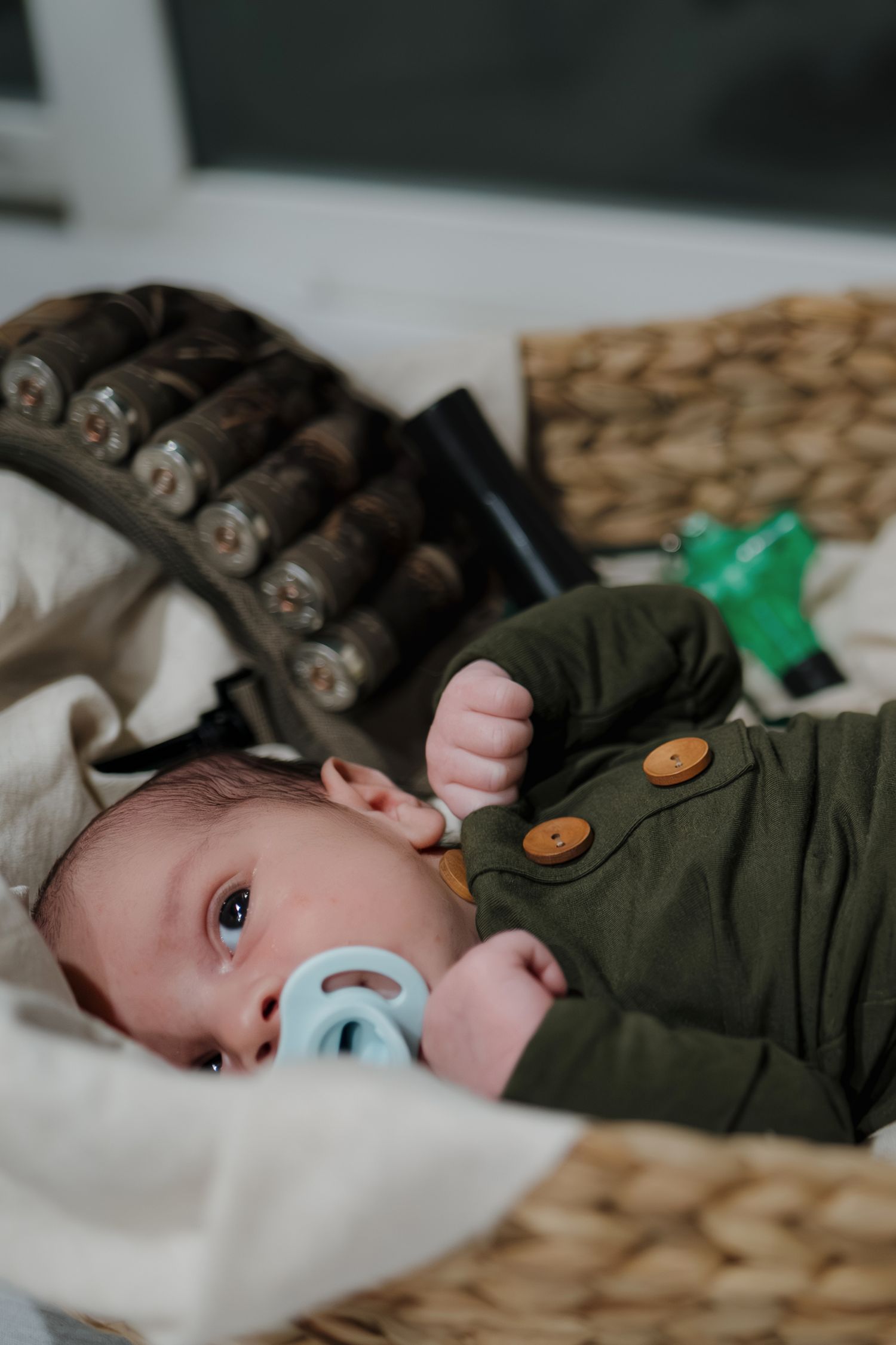 A small baby wearing a dark green outfit with wooden buttons lies on white bedding with a blue pacifier.