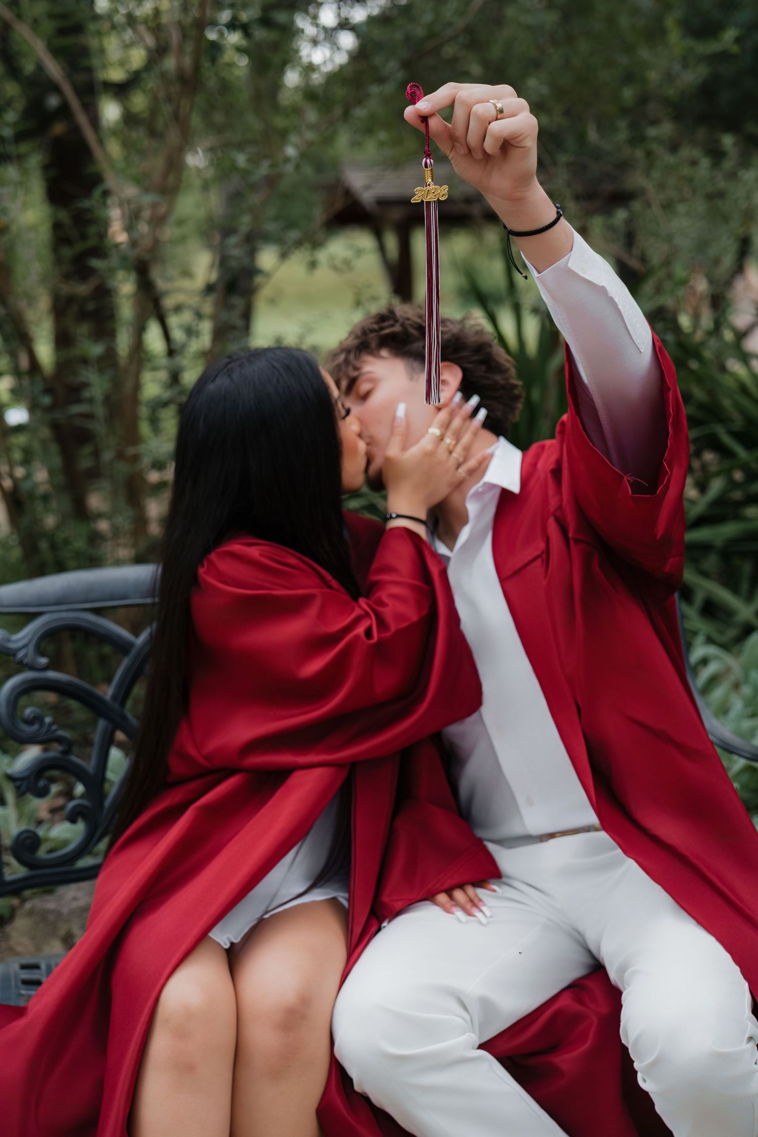 Two graduates in red robes share a kiss on a bench while holding a diploma, surrounded by lush greenery.