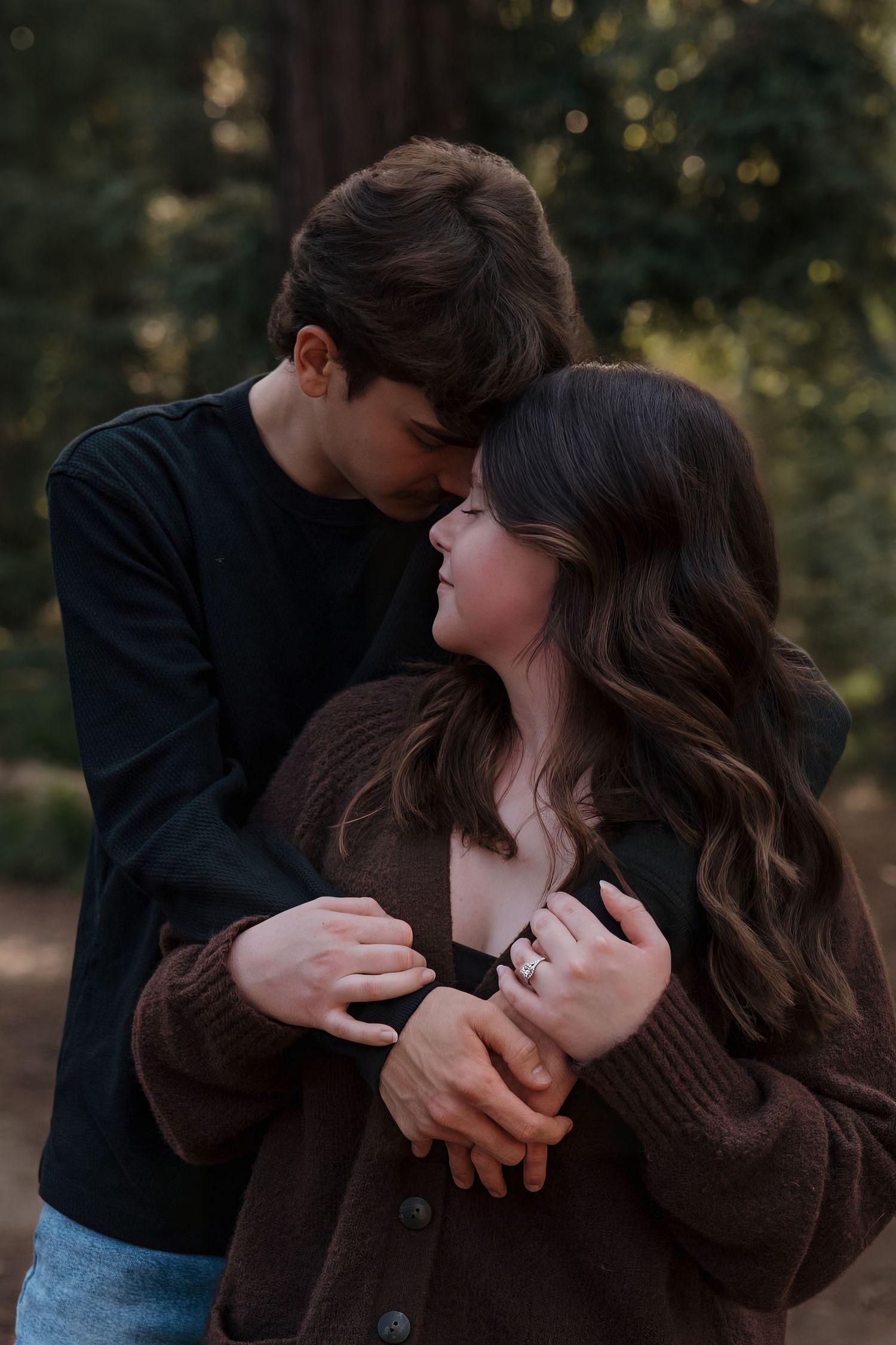 A couple shares a tender embrace in a forest, the woman smiling up as the man leans in close, both in dark cozy outfits.