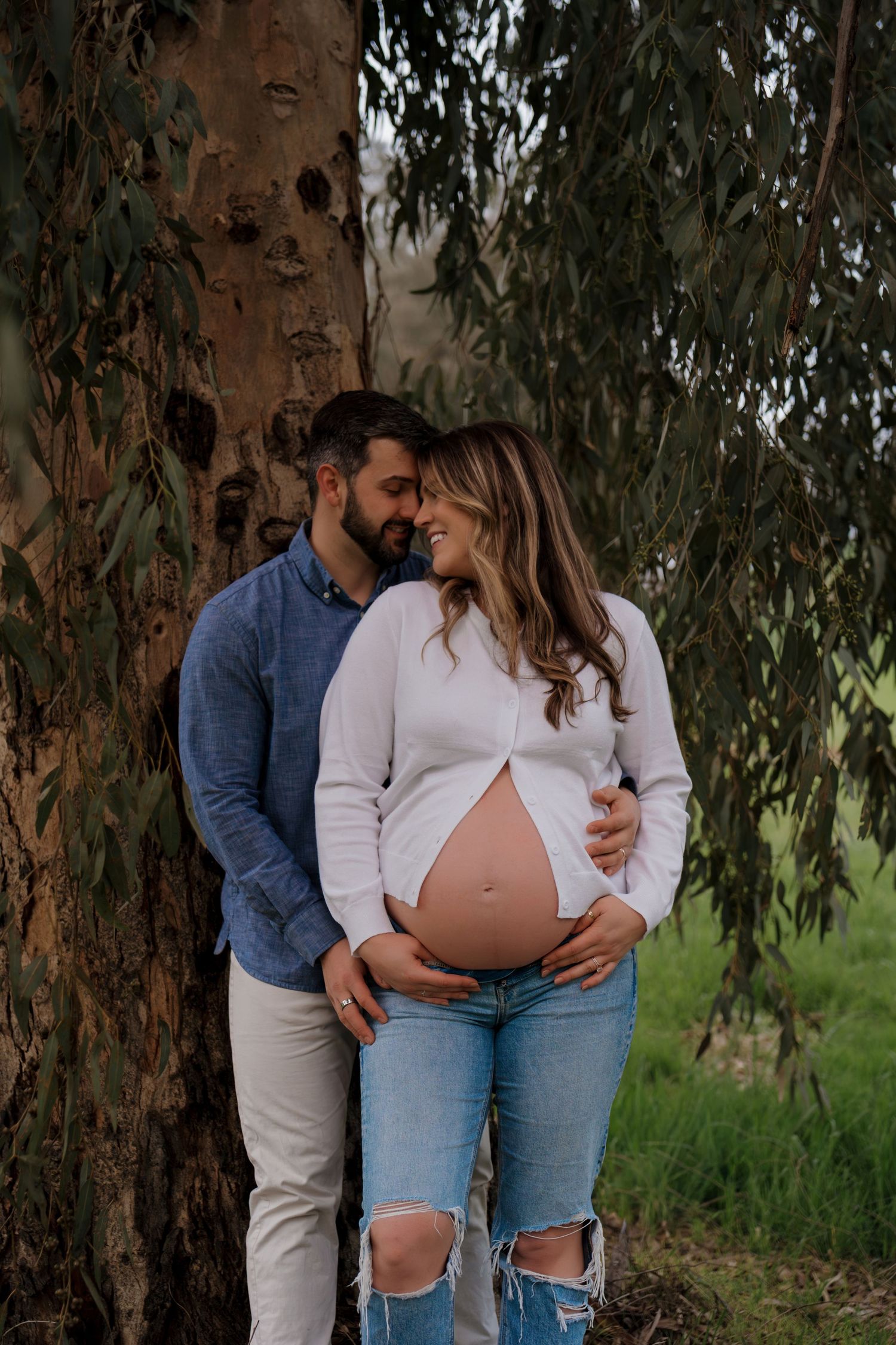 An expectant couple shares tender moments outdoors near a tree trunk, wearing coordinated casual outfits.