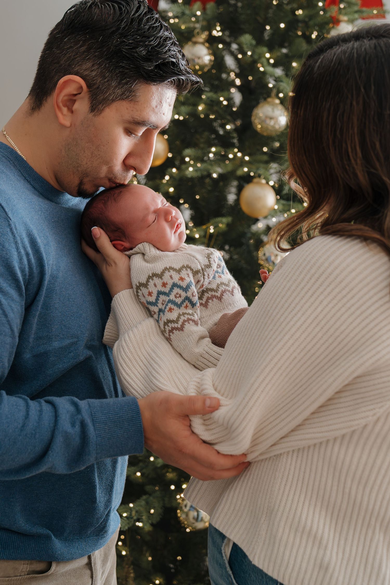 A couple shares a tender moment in front of a Christmas tree decorated with white ornaments and twinkling lights.