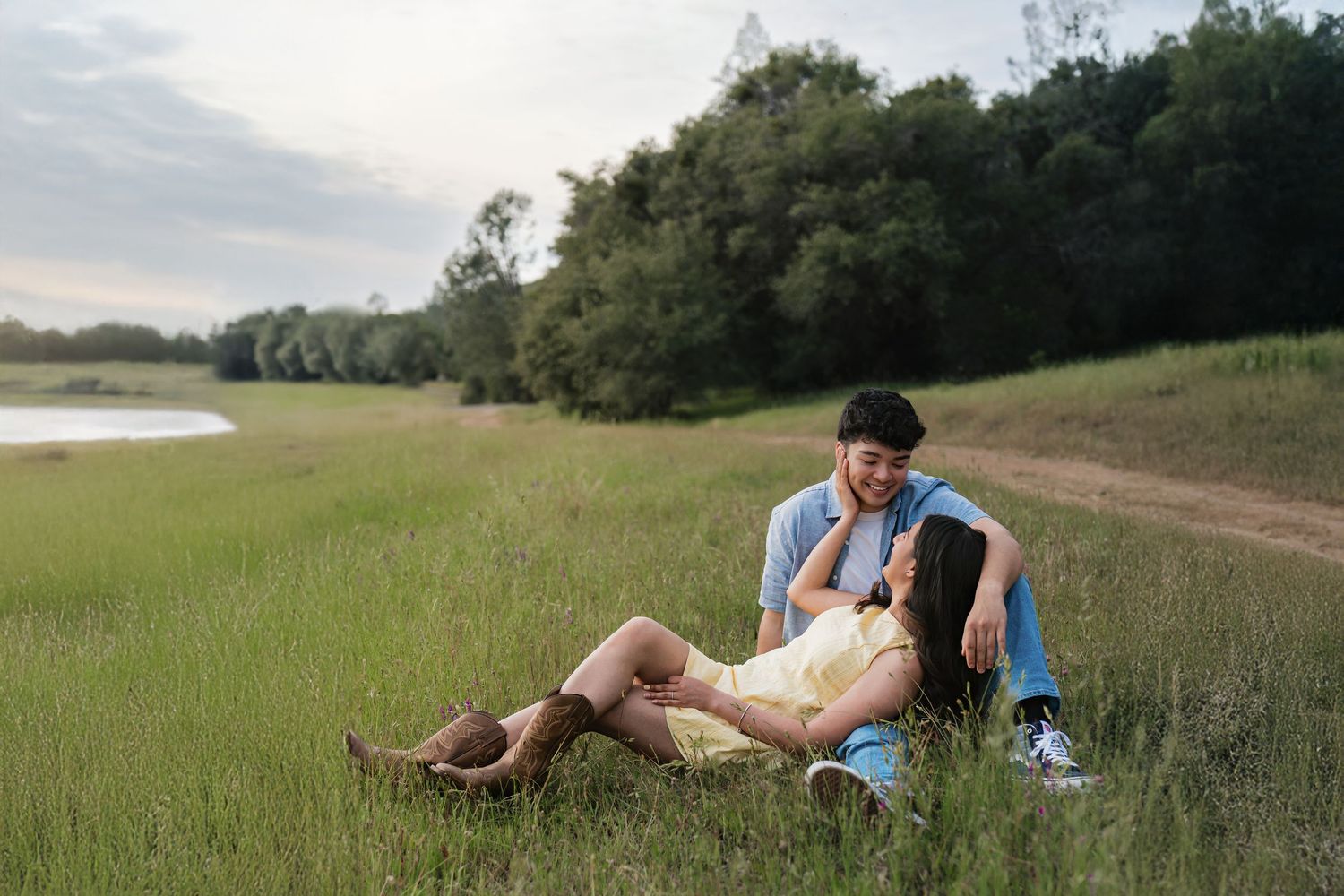 Couple sitting together in a lush green meadow near a lake, sharing a romantic moment outdoors.