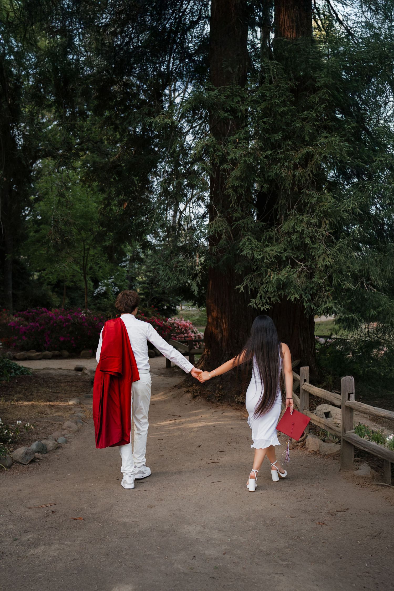 Couple in white outfits holding hands walking through a scenic tree-lined park path.