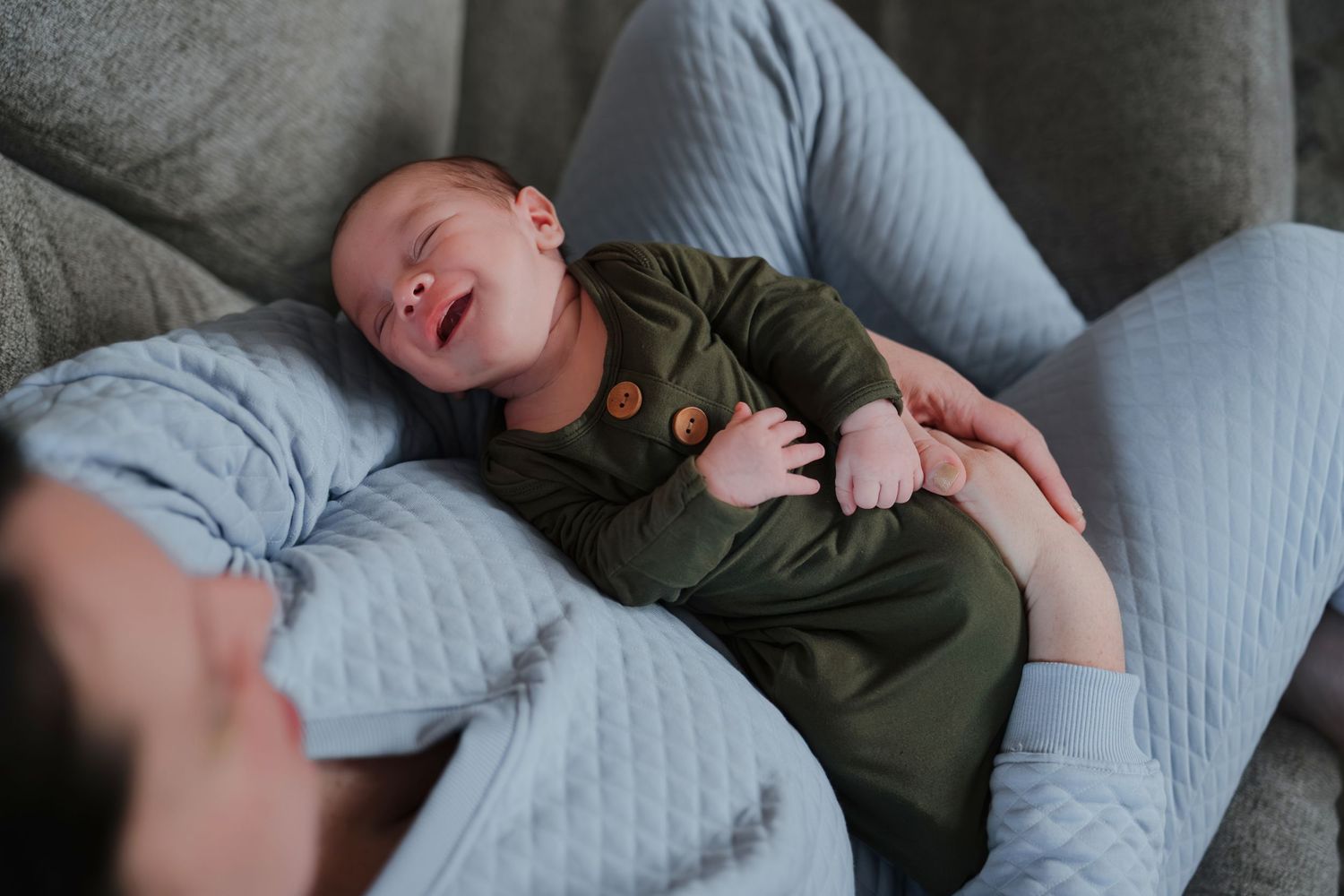 Sleeping baby wearing olive green outfit rests peacefully on quilted gray surface while being comforted.