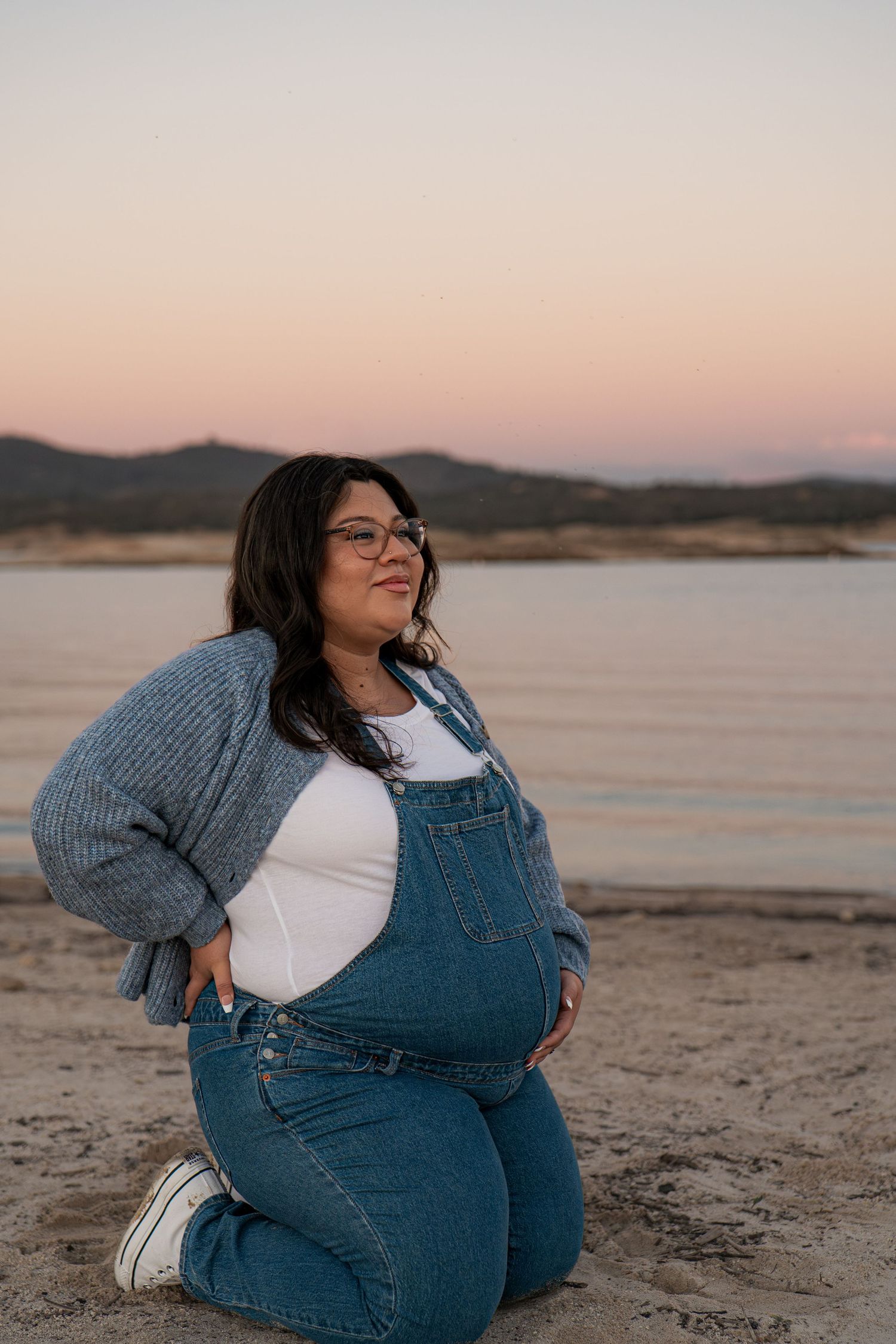 Person in denim overalls sits on beach at sunset with mountains in background in Roseville, CA