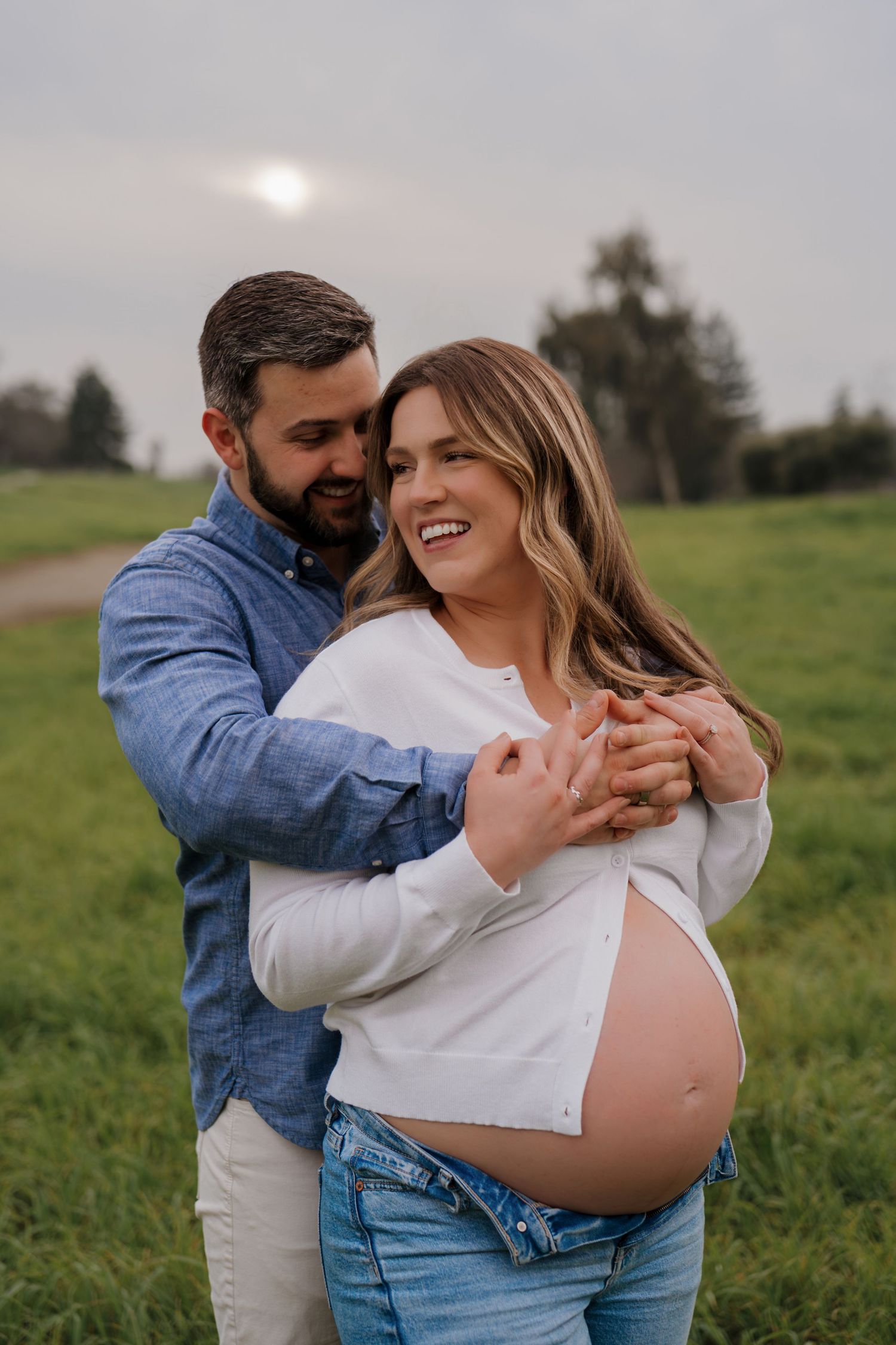 A couple embraces in a grassy field during a romantic maternity photoshoot at sunset.