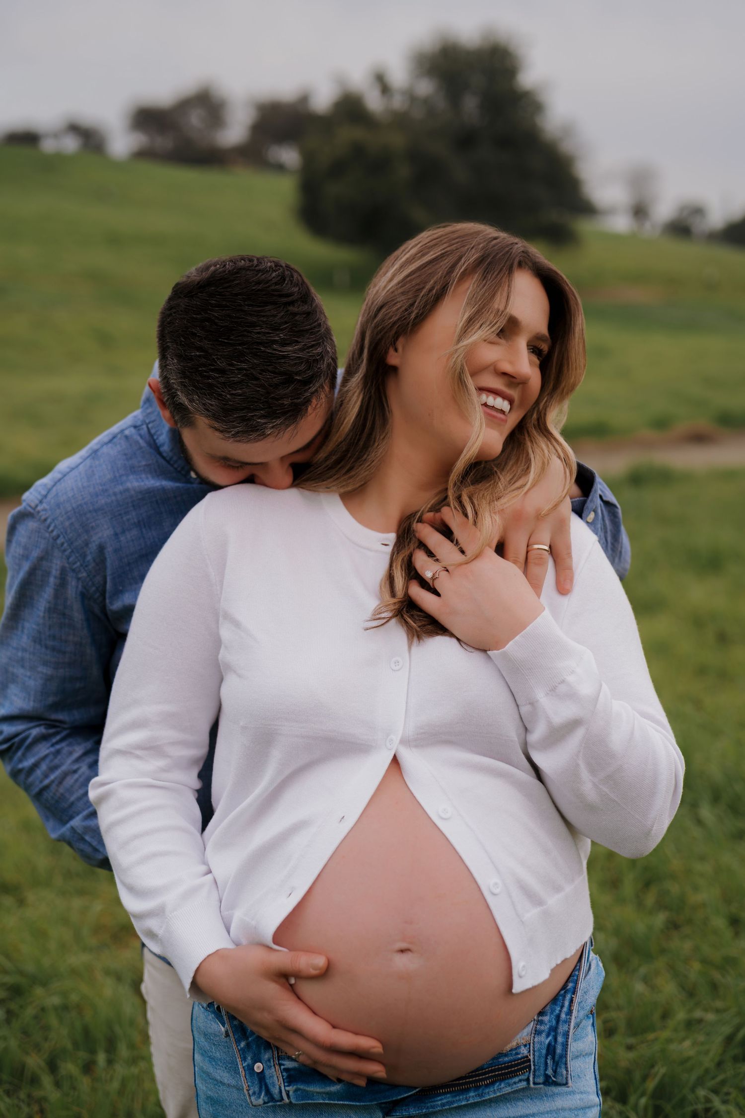 Couple shares intimate moment in grassy field during maternity photoshoot outdoors.