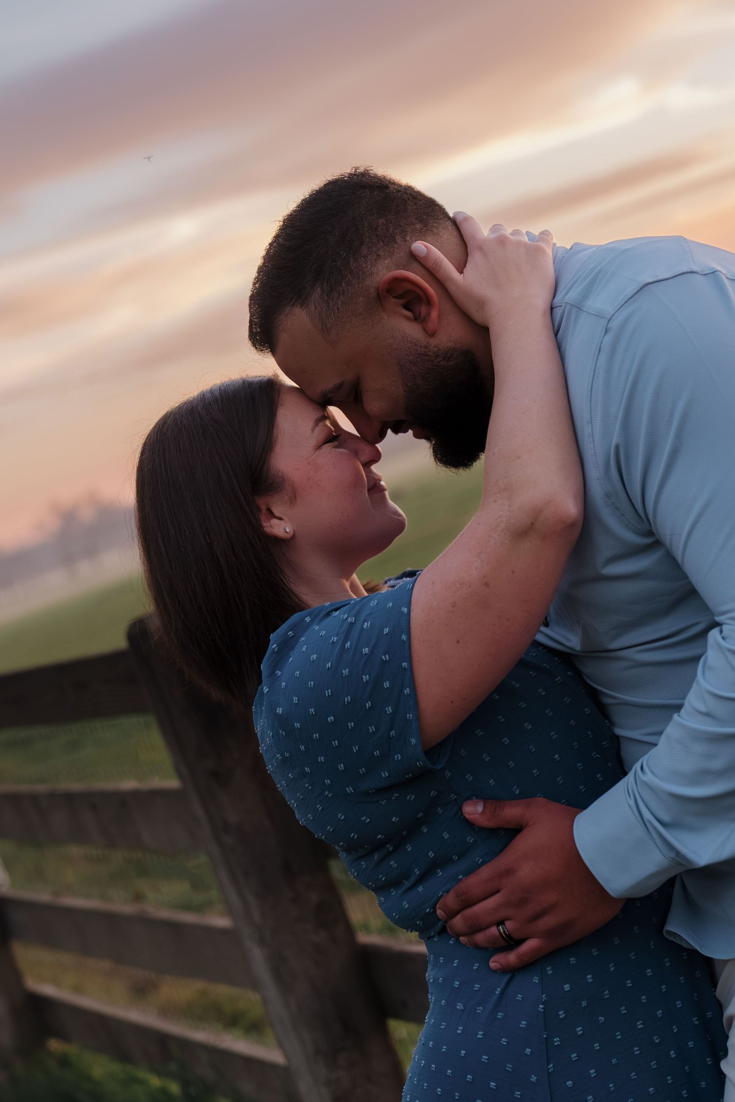 A romantic couple in a blue dress and gray shirt embrace against a sunset backdrop in a rural landscape.