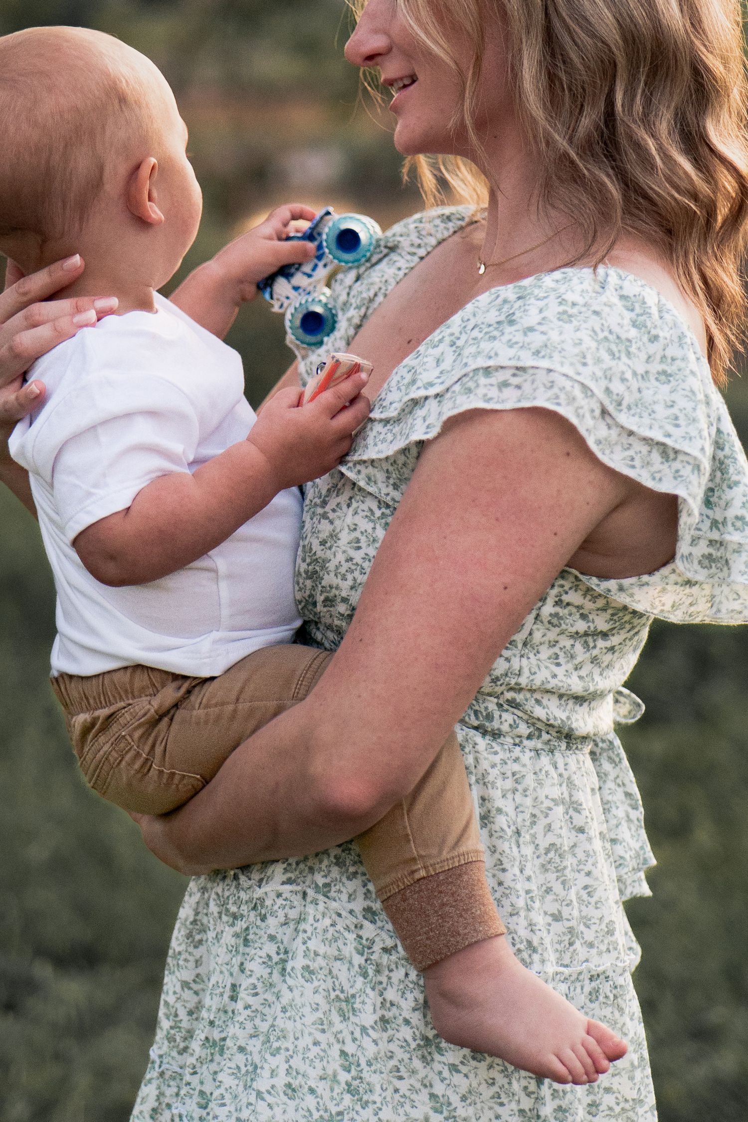 Parent holding baby in white onesie against floral dress backdrop outdoors.