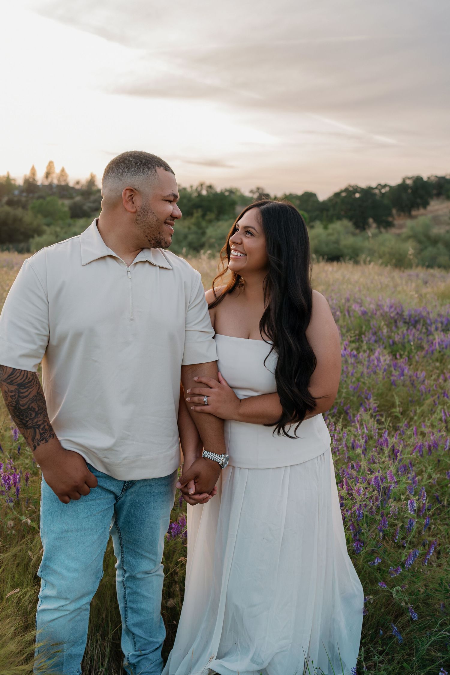 A couple smiles at each other in a purple wildflower field at sunset, woman in white dress, man in white polo and jeans.