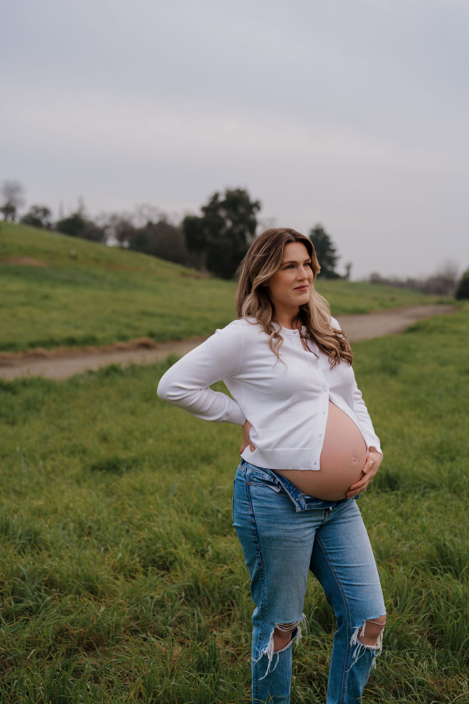 A maternity photo session in a grassy field shows a woman in ripped jeans and white top cradling her baby bump.