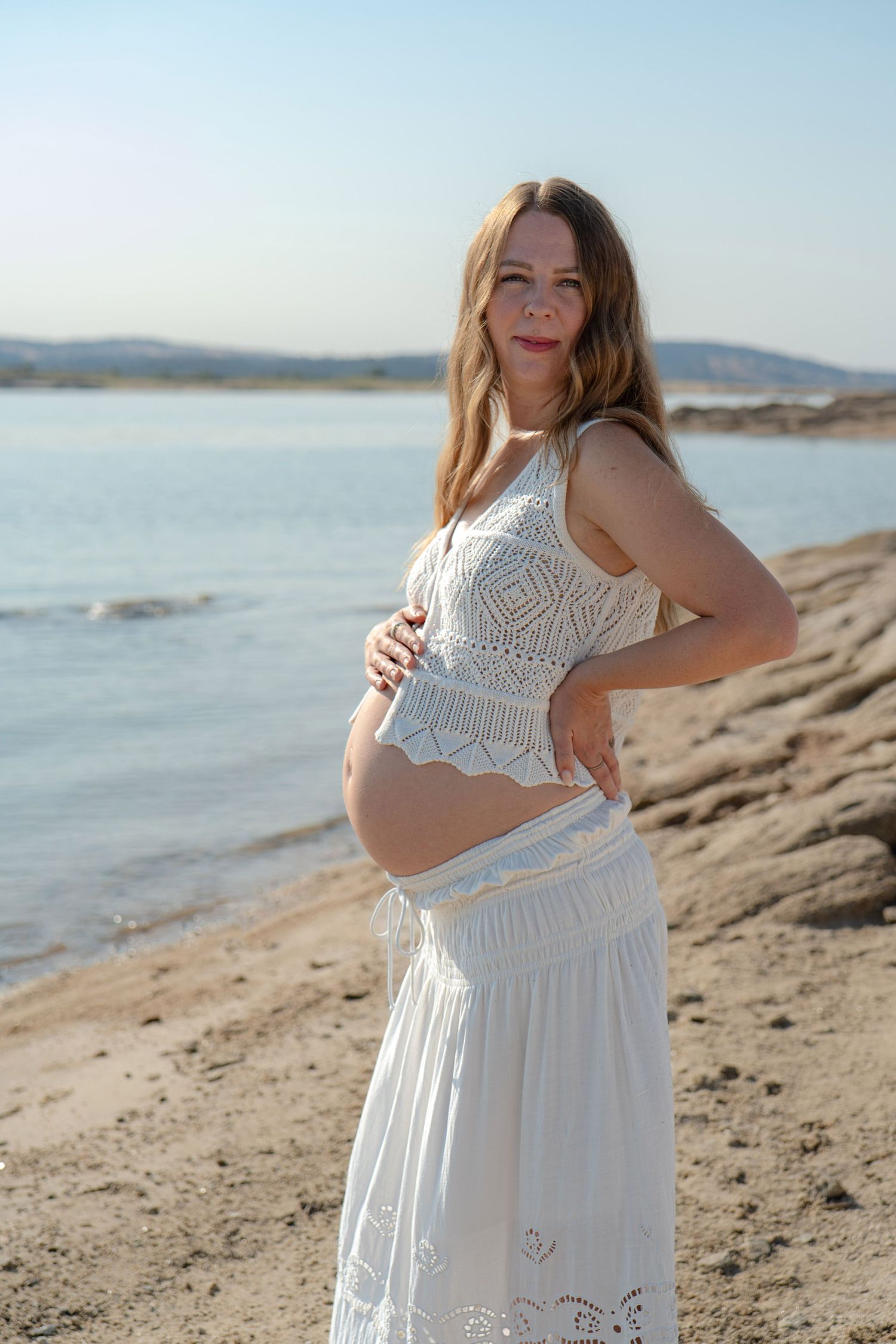 Person in white lace dress poses on rocky beach with mountains in background in Roseville, CA