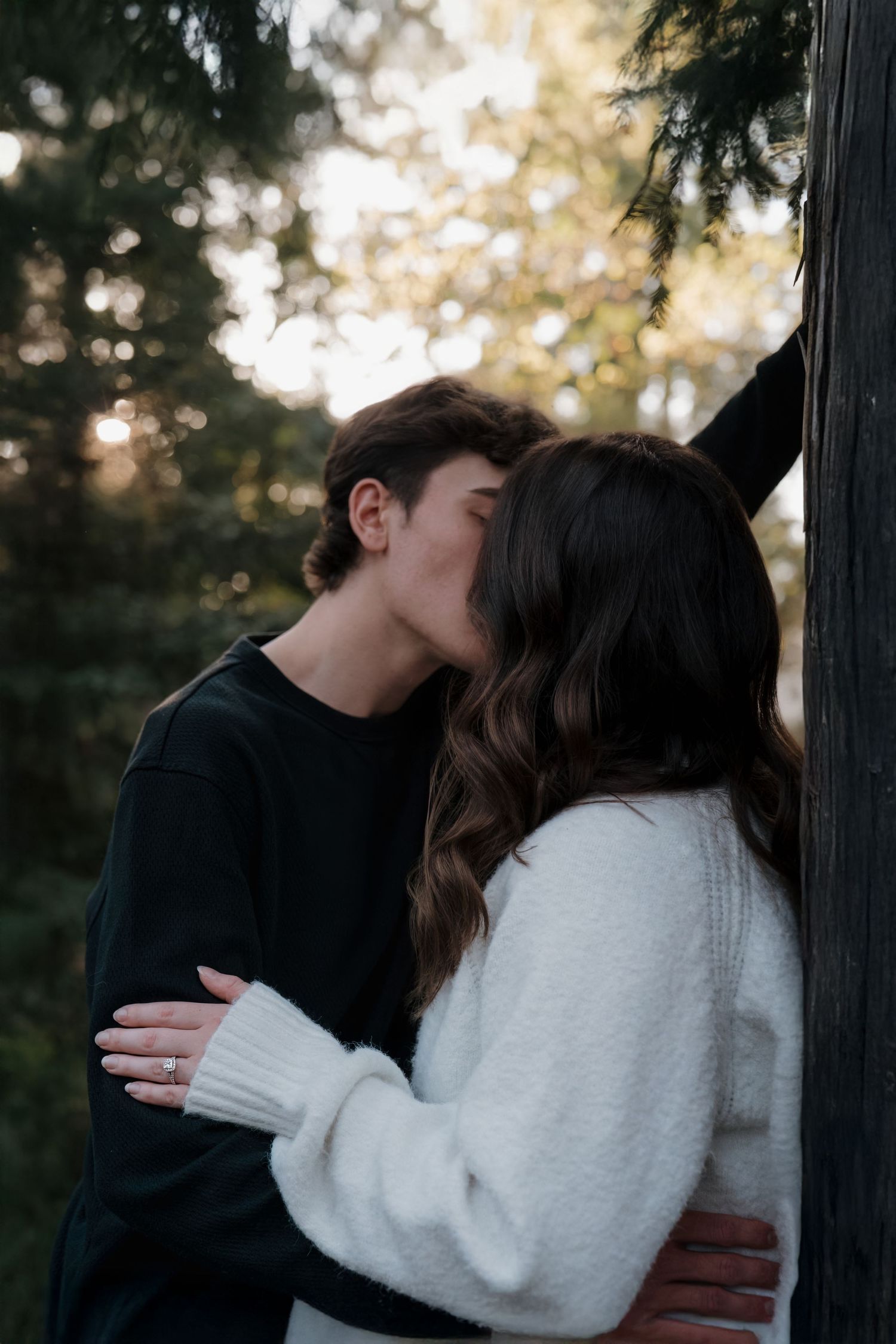 A couple shares a romantic kiss outdoors near a tree, surrounded by lush green forest in soft golden light.