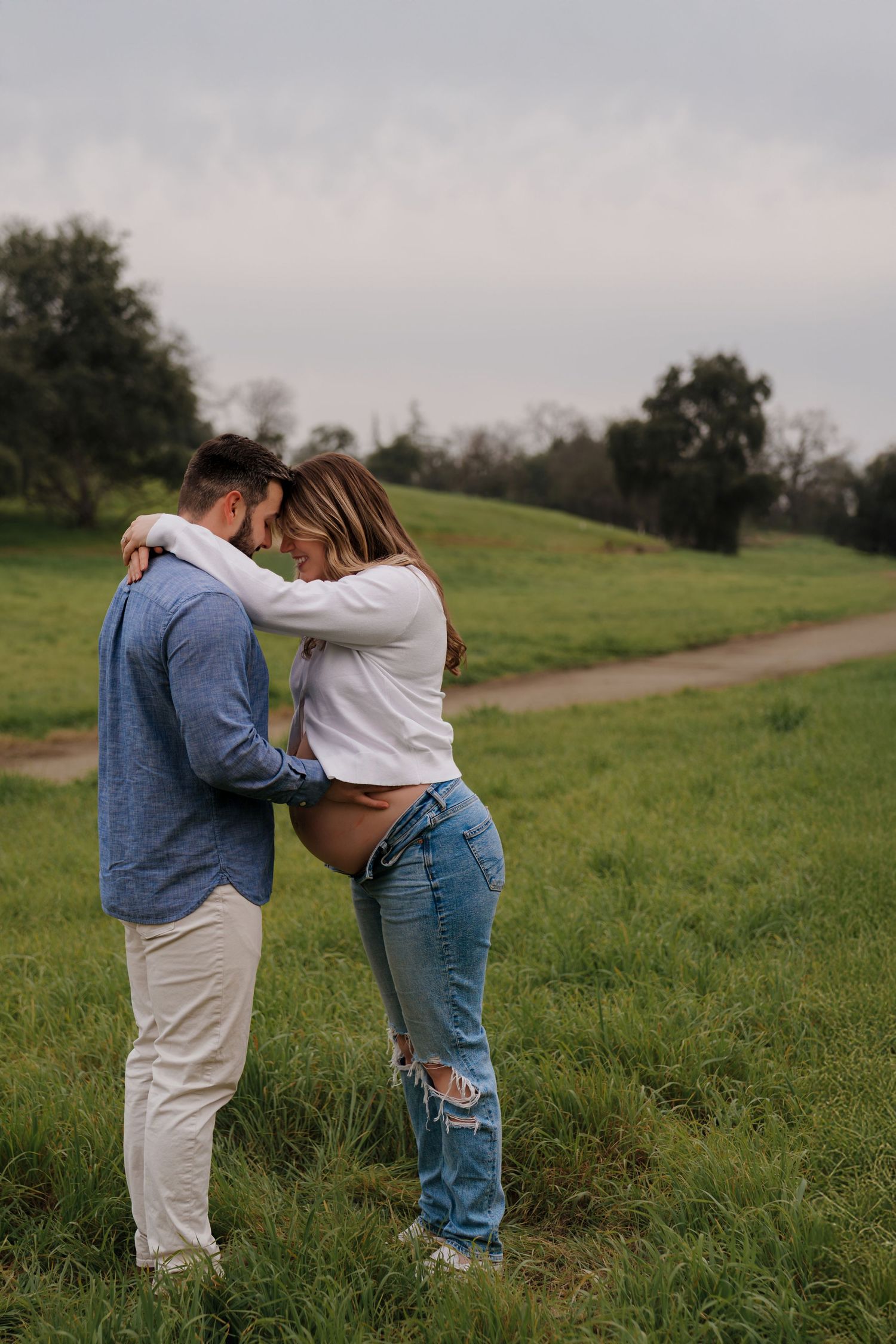 A couple embraces tenderly in a grassy field on an overcast day, wearing casual denim and white clothing.