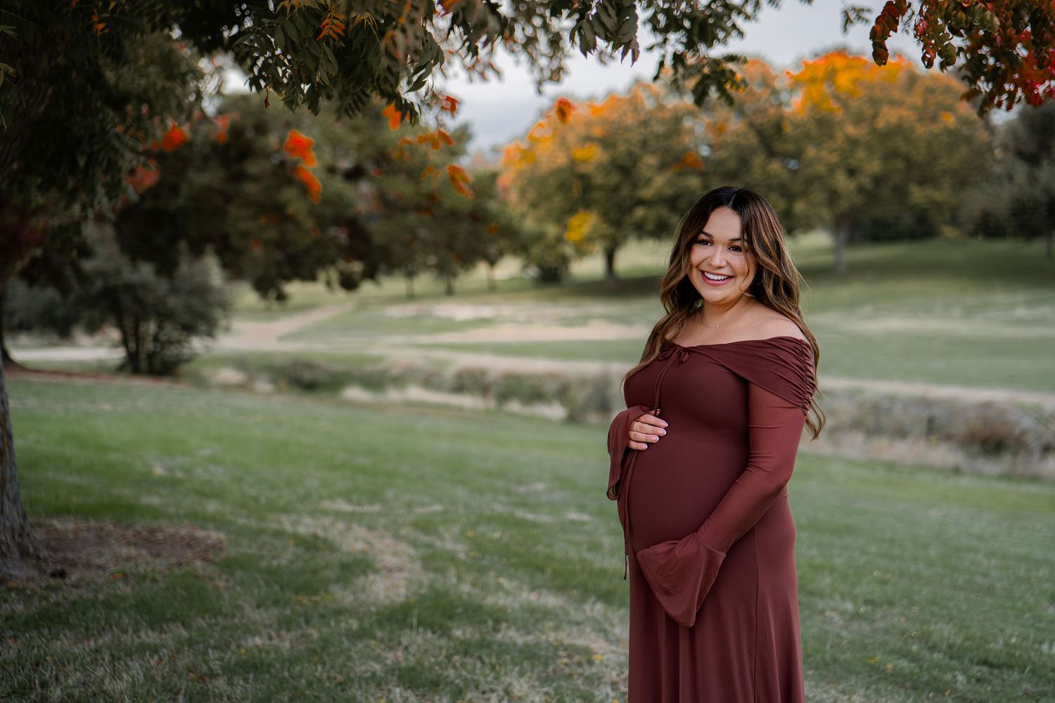 Autumn maternity photoshoot in burgundy dress among fall foliage near lake in Roseville, CA