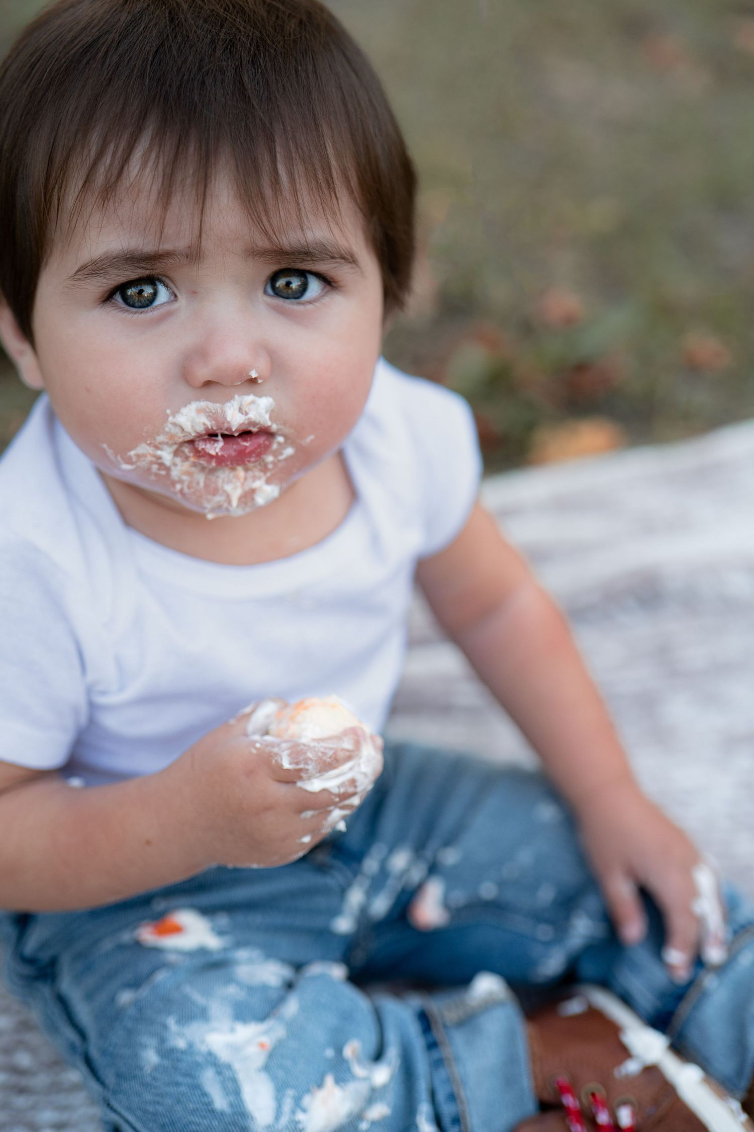 A small child in a white shirt and jeans enjoys birthday cake with messy frosting all over their face and nose.