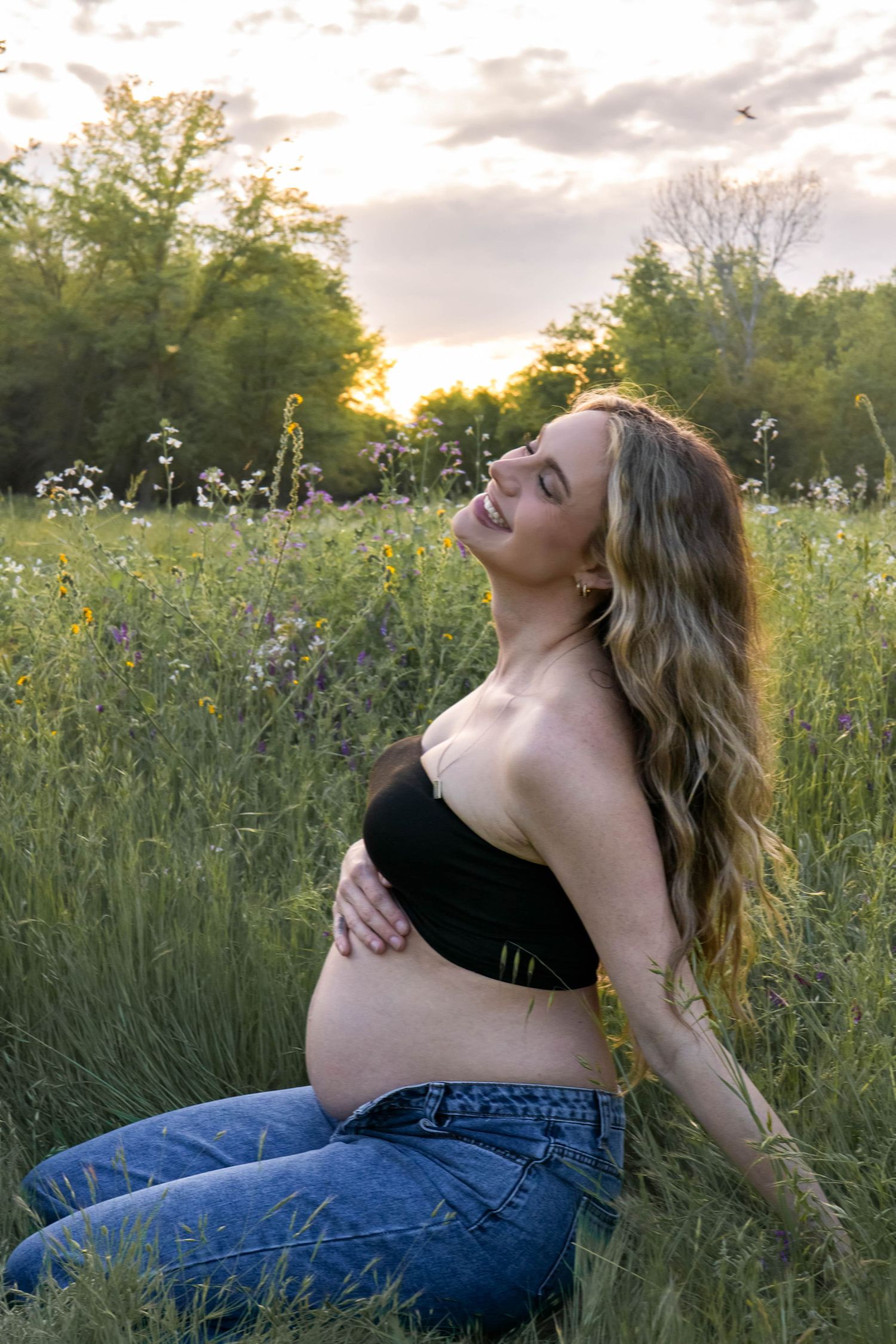 Pregnant woman in black top and jeans reclines in wildflower meadow at sunset.