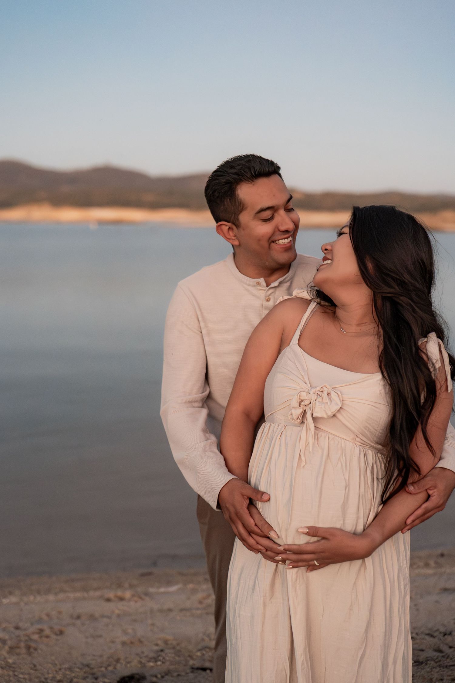 Couple in light colored outfits embracing on beach at sunset.