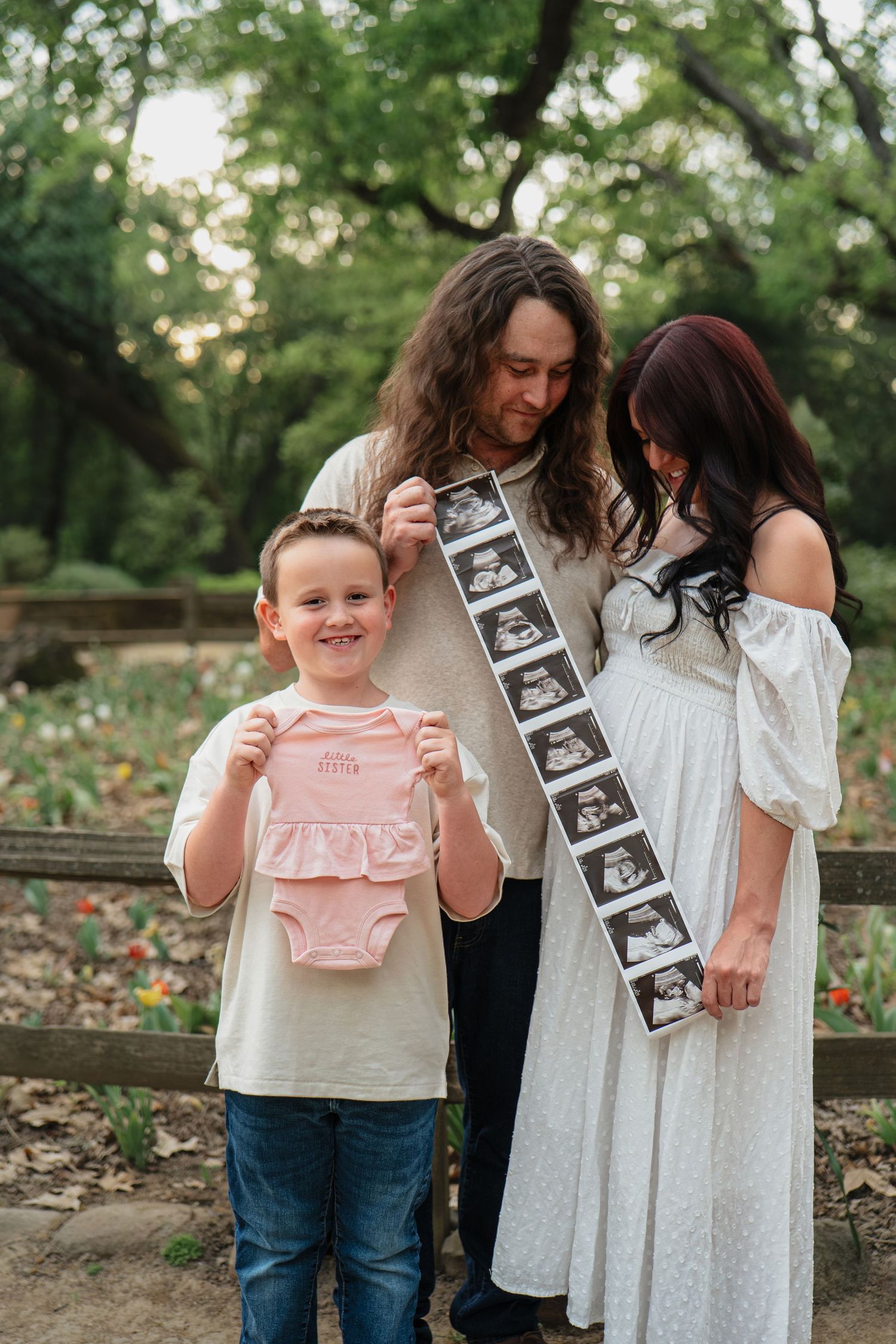 Family pregnancy announcement outdoors, child holding pink onesie, parents displaying ultrasound strip, garden setting.