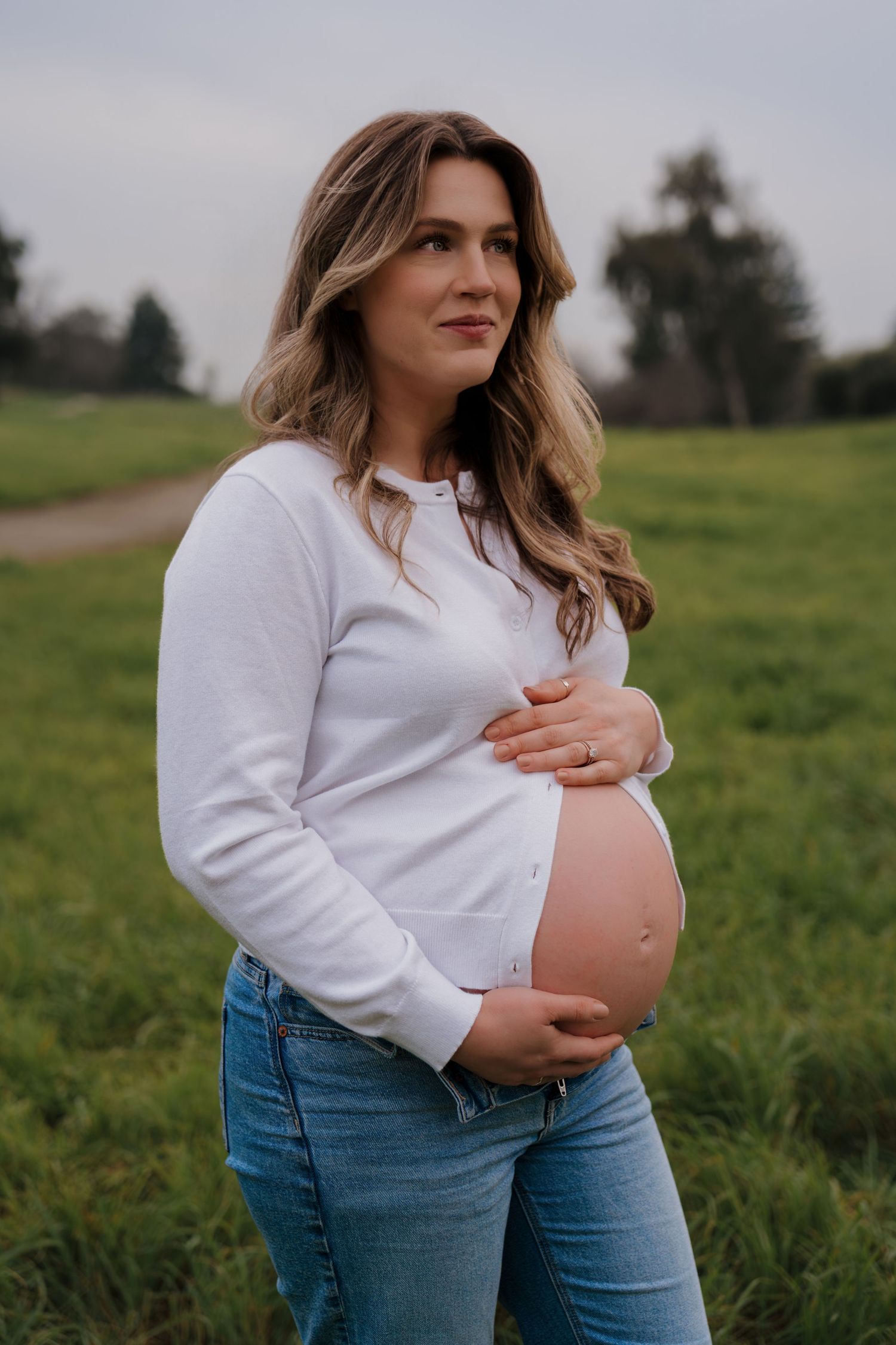 A series of maternity photos showing a pregnant person in a white sweater and jeans standing in a green field.