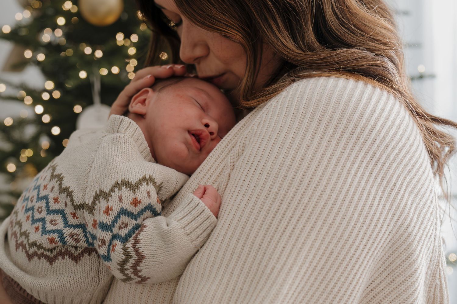 A sleeping baby in a patterned sweater rests against someone wearing a cream knit sweater with a Christmas tree in background.