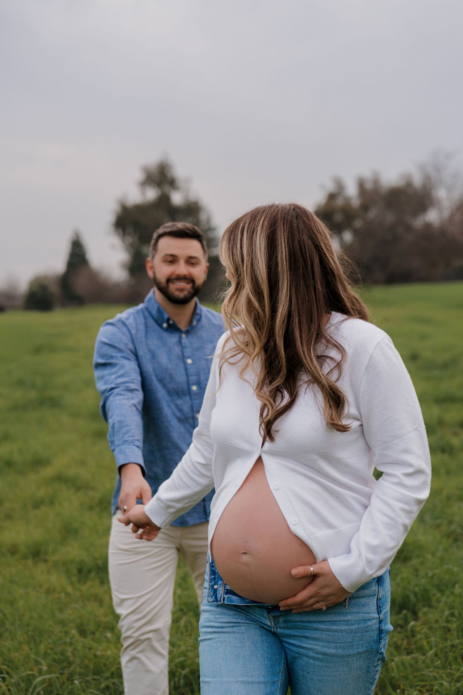 A couple shares playful moments in a green field during a maternity photoshoot wearing white and denim outfits.