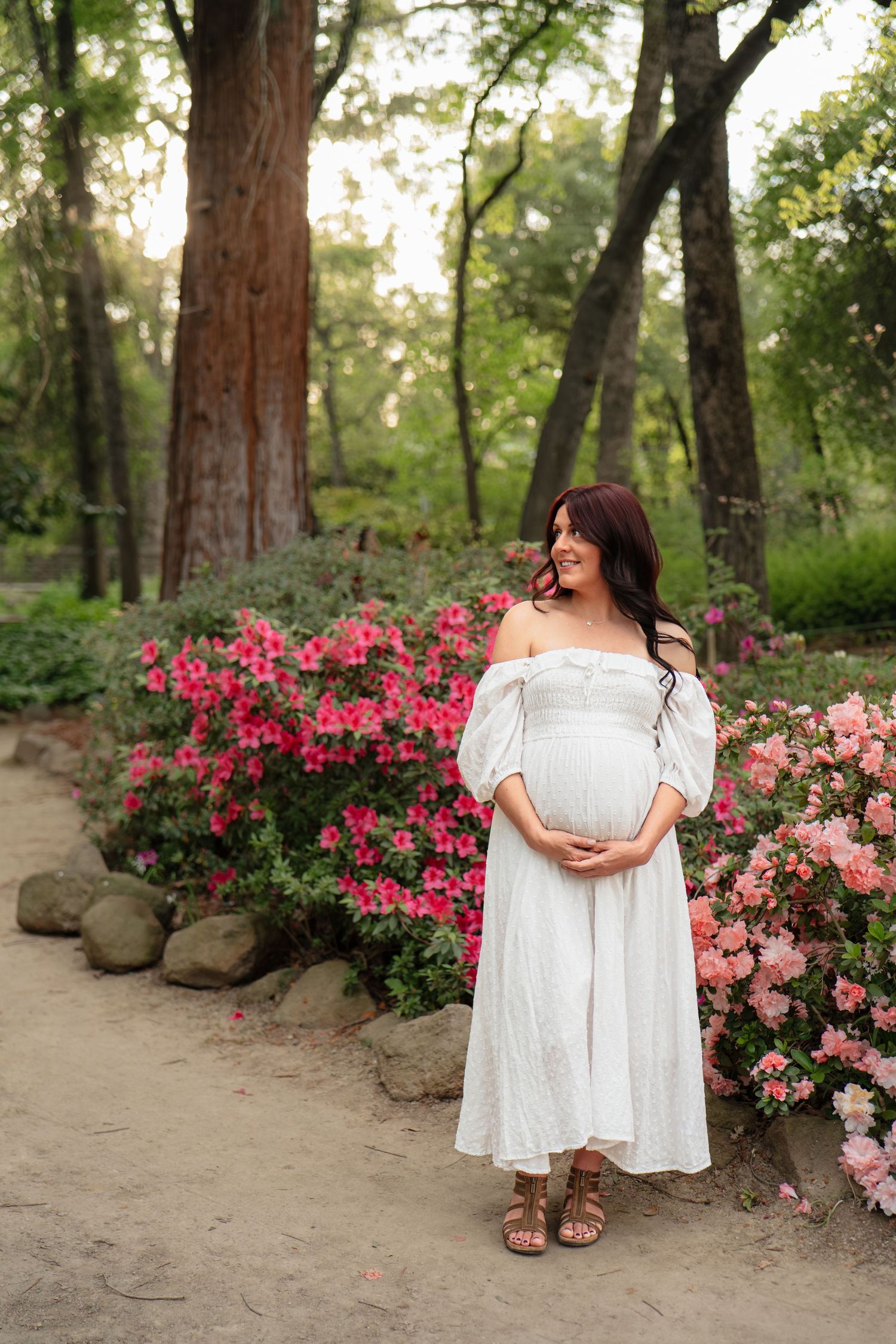 Pregnant woman in white off-shoulder dress holding baby bump, standing among pink flowering bushes in a lush garden park.