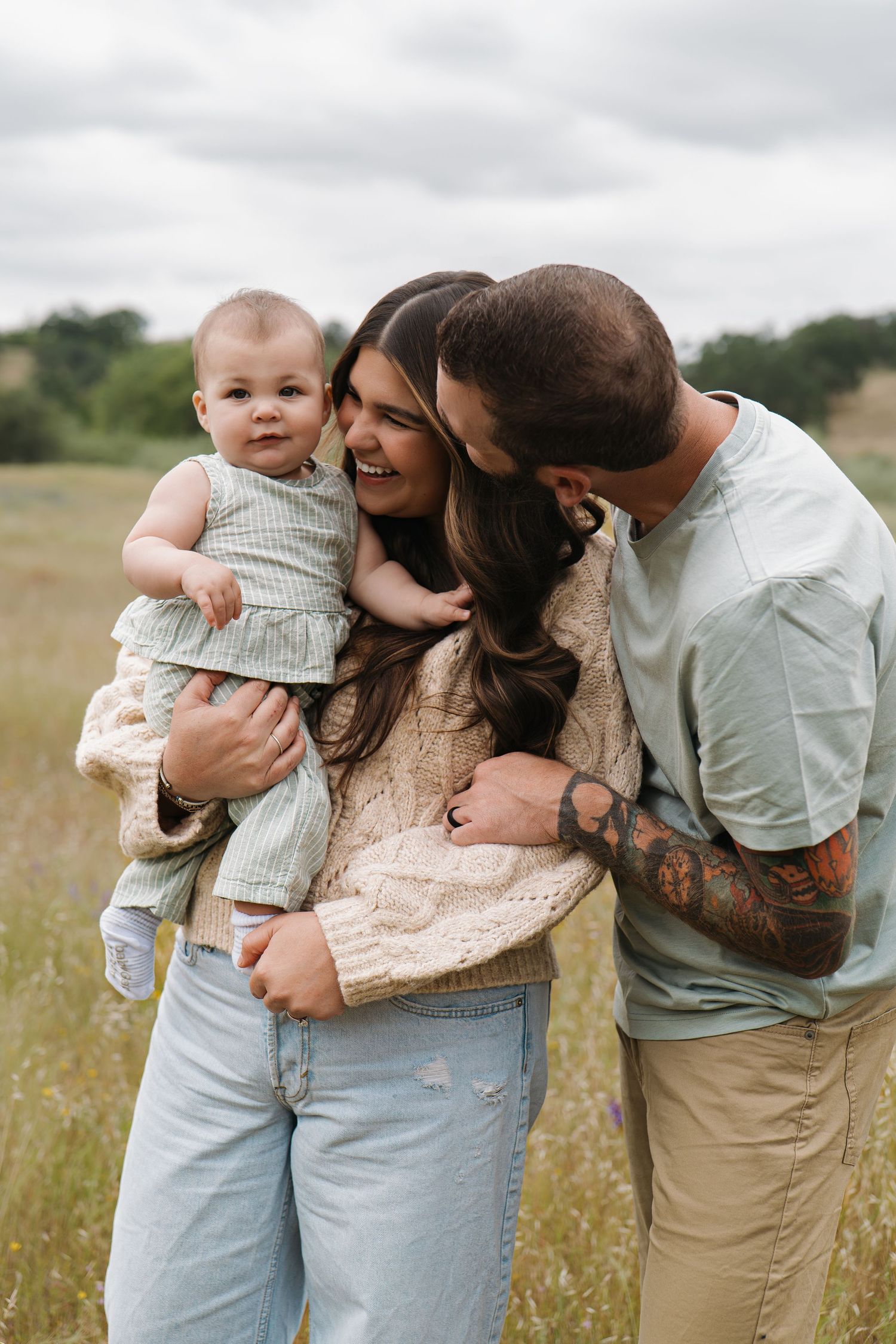 A happy young family with a tattooed dad kissing mom while she holds their baby in a scenic outdoor field.