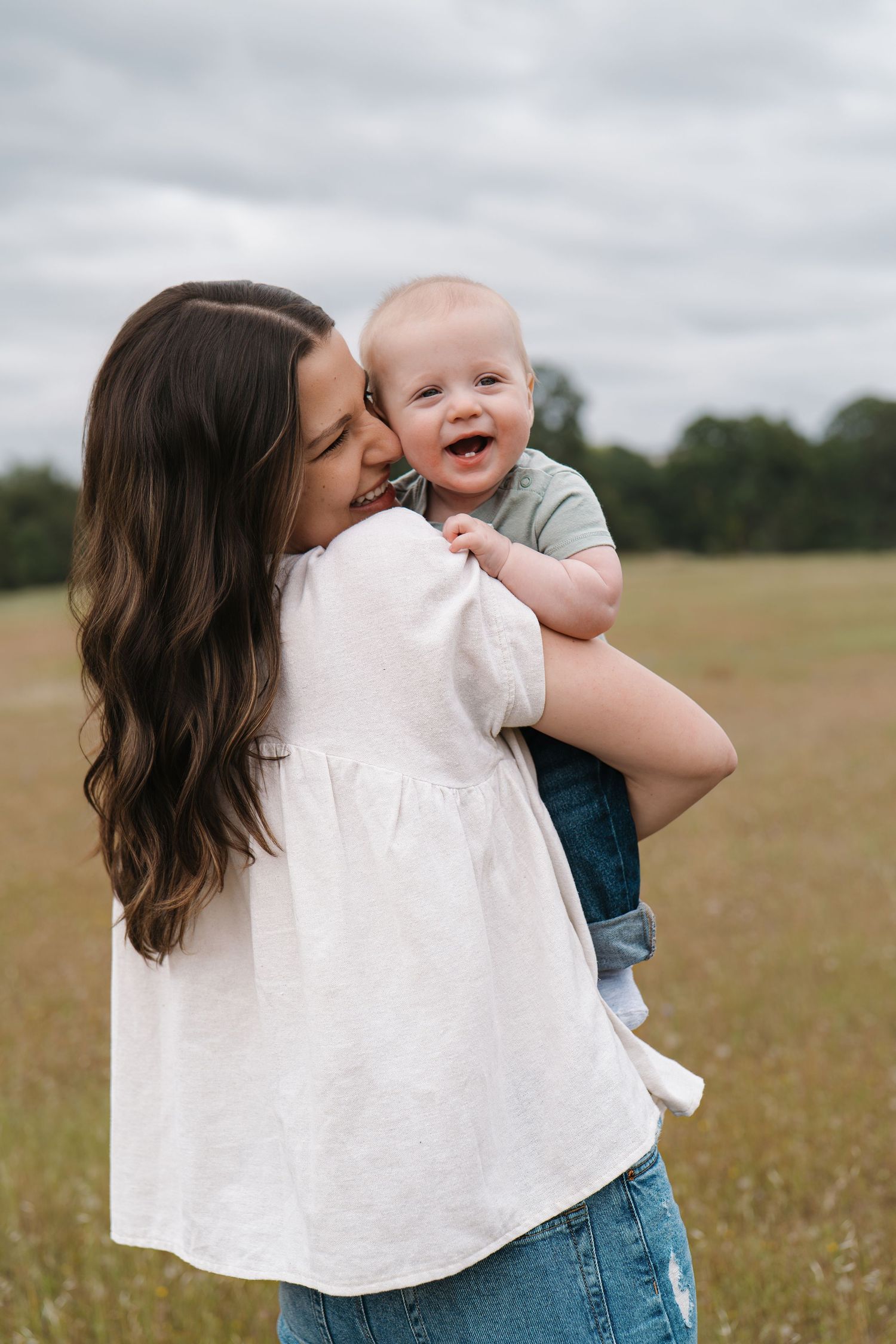 A mother with long dark hair kisses her laughing baby outdoors in a green field.