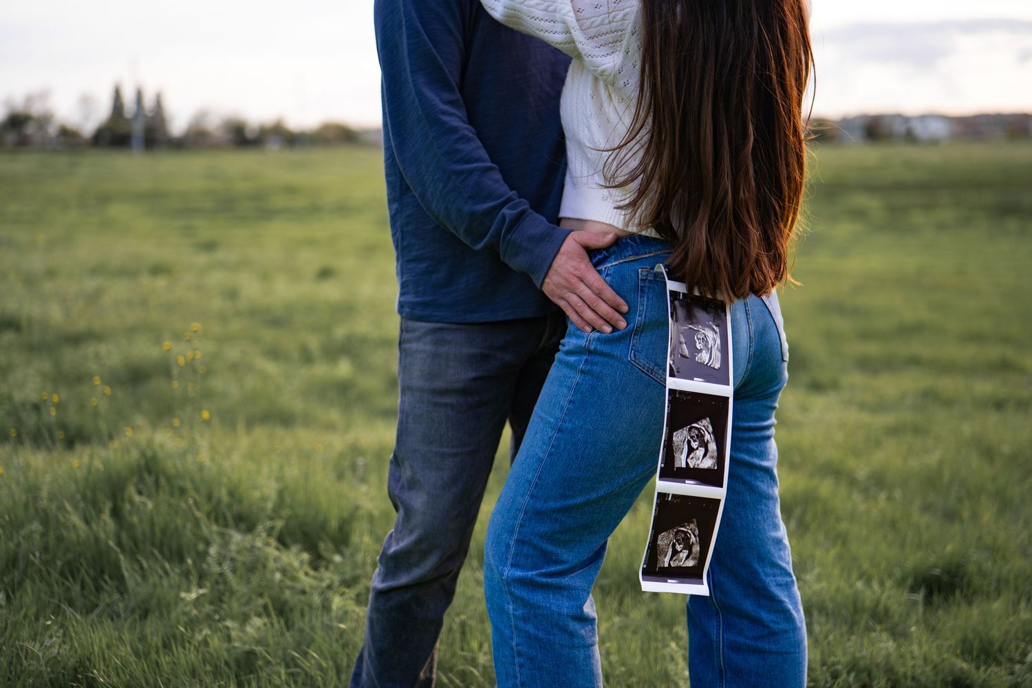 Couple holding ultrasound photos in a grassy field during golden hour sunset.