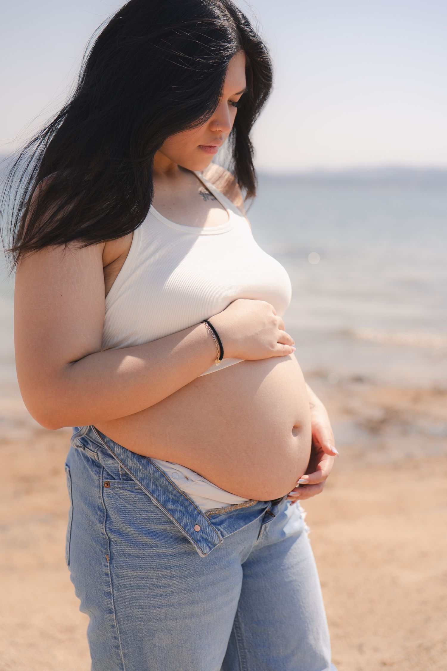 Maternity photos at the beach showing a growing baby bump in casual white top and jeans.