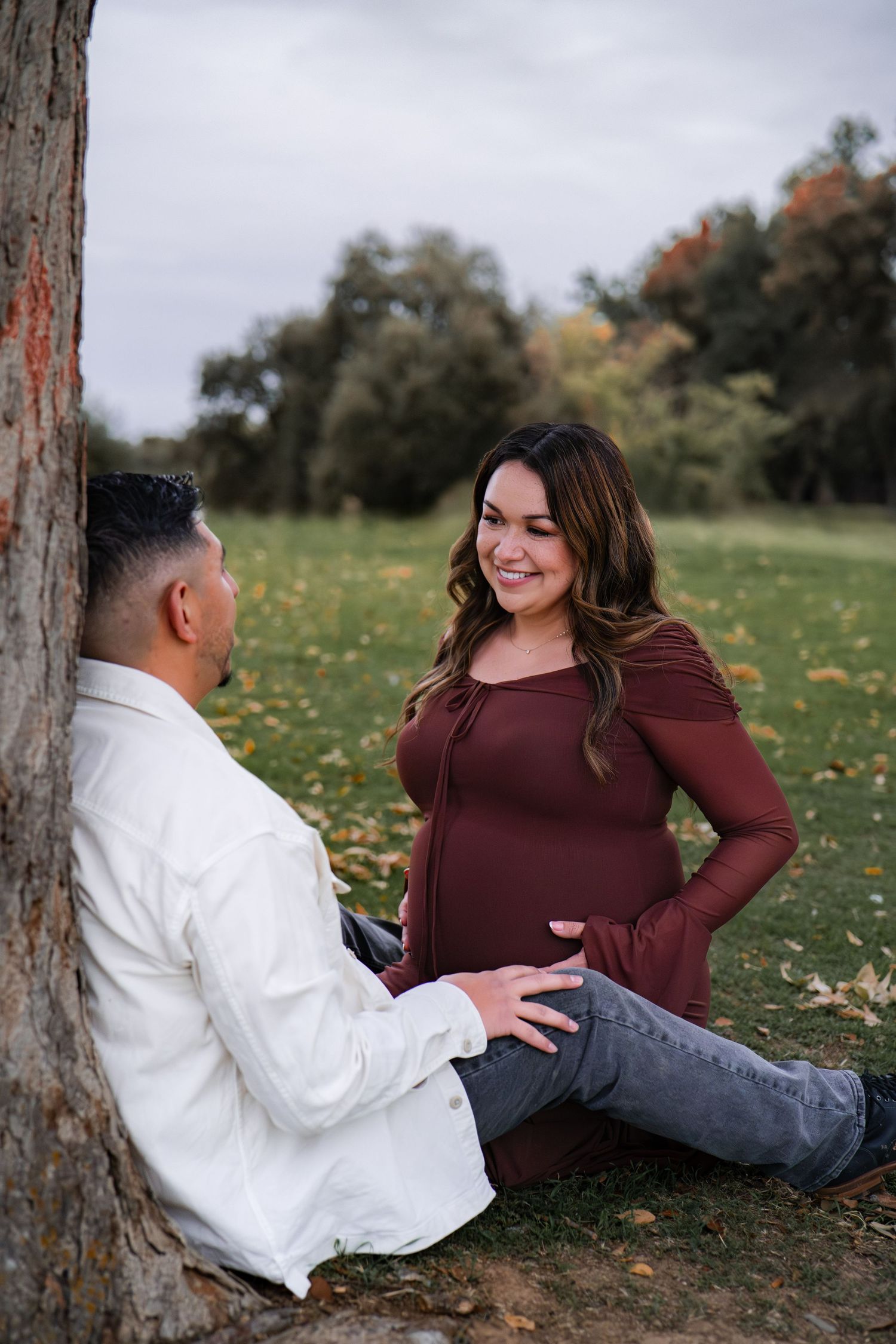 A couple sits together on grass during an autumn photoshoot in a park setting.