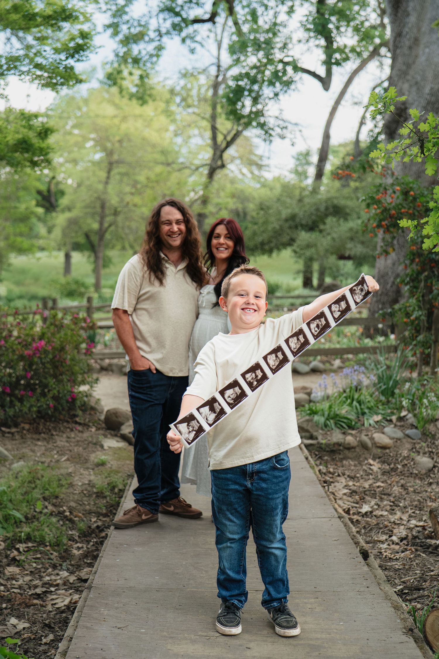 Happy family maternity announcement with young boy proudly displaying ultrasound photos on a garden pathway with parents smiling behind him.