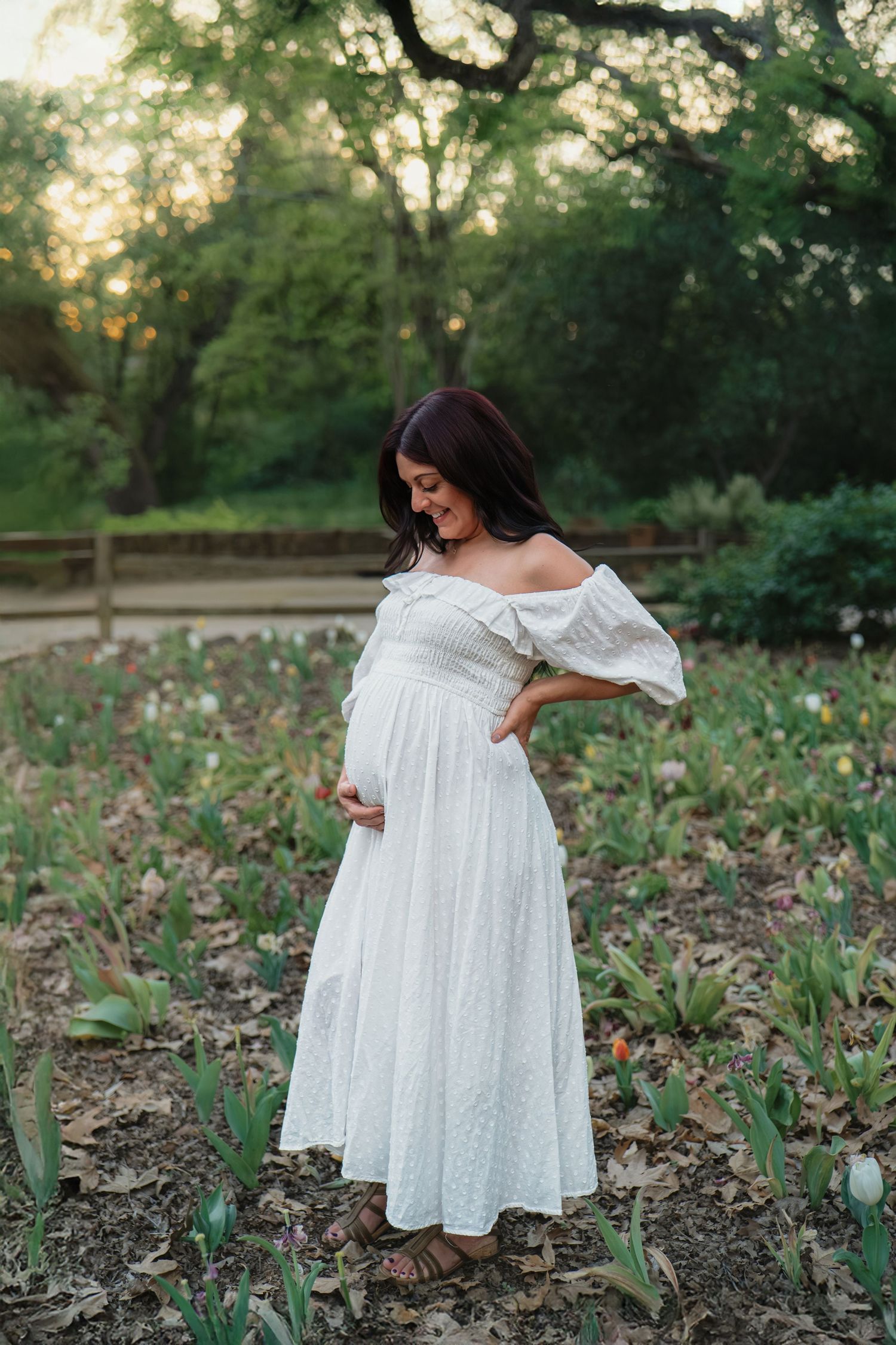 Pregnant woman in white off-shoulder dress cradling baby bump in a garden with tulips and trees at sunset.