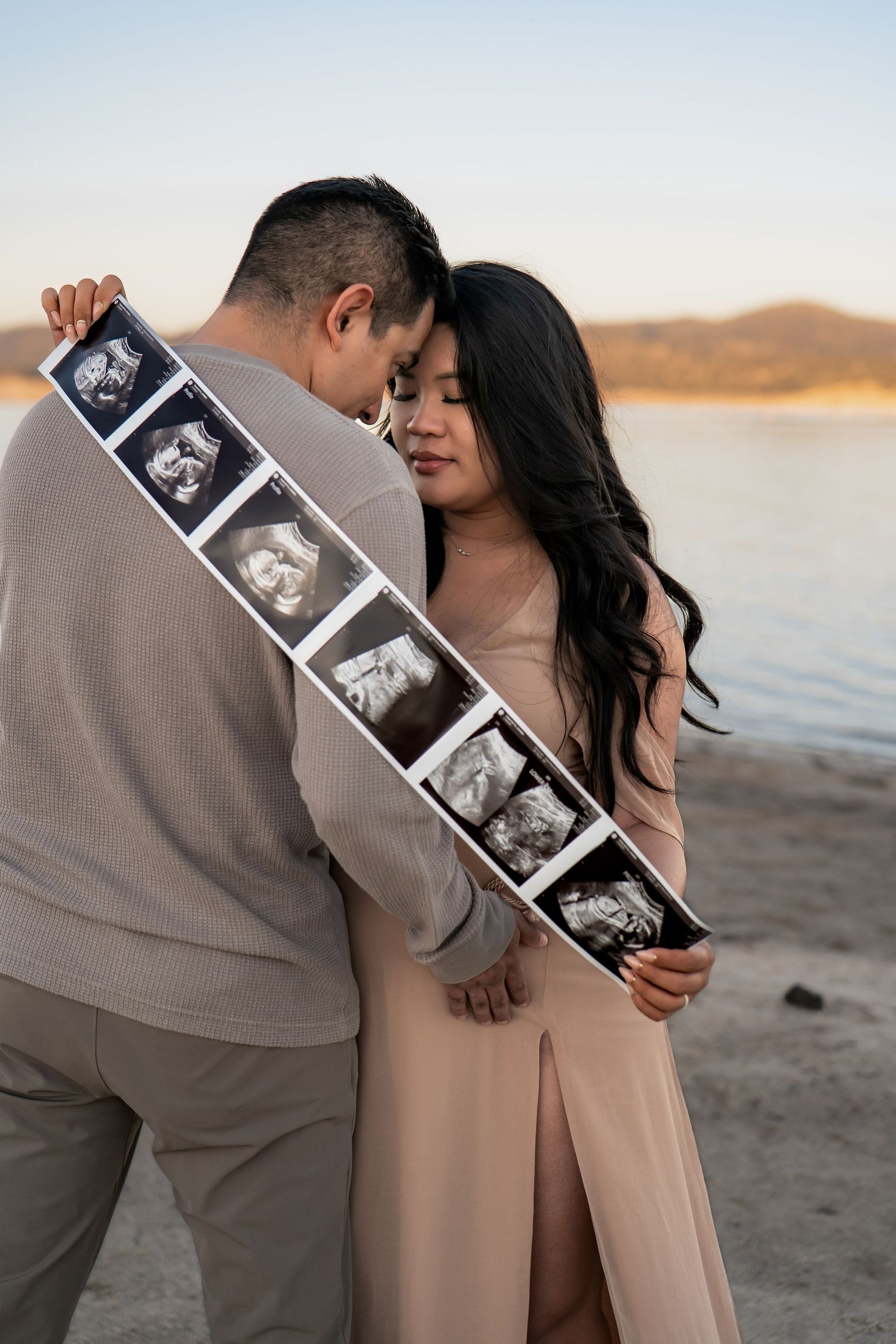Couple sharing intimate moments on a beach at sunset while holding ultrasound photos in Roseville, CA