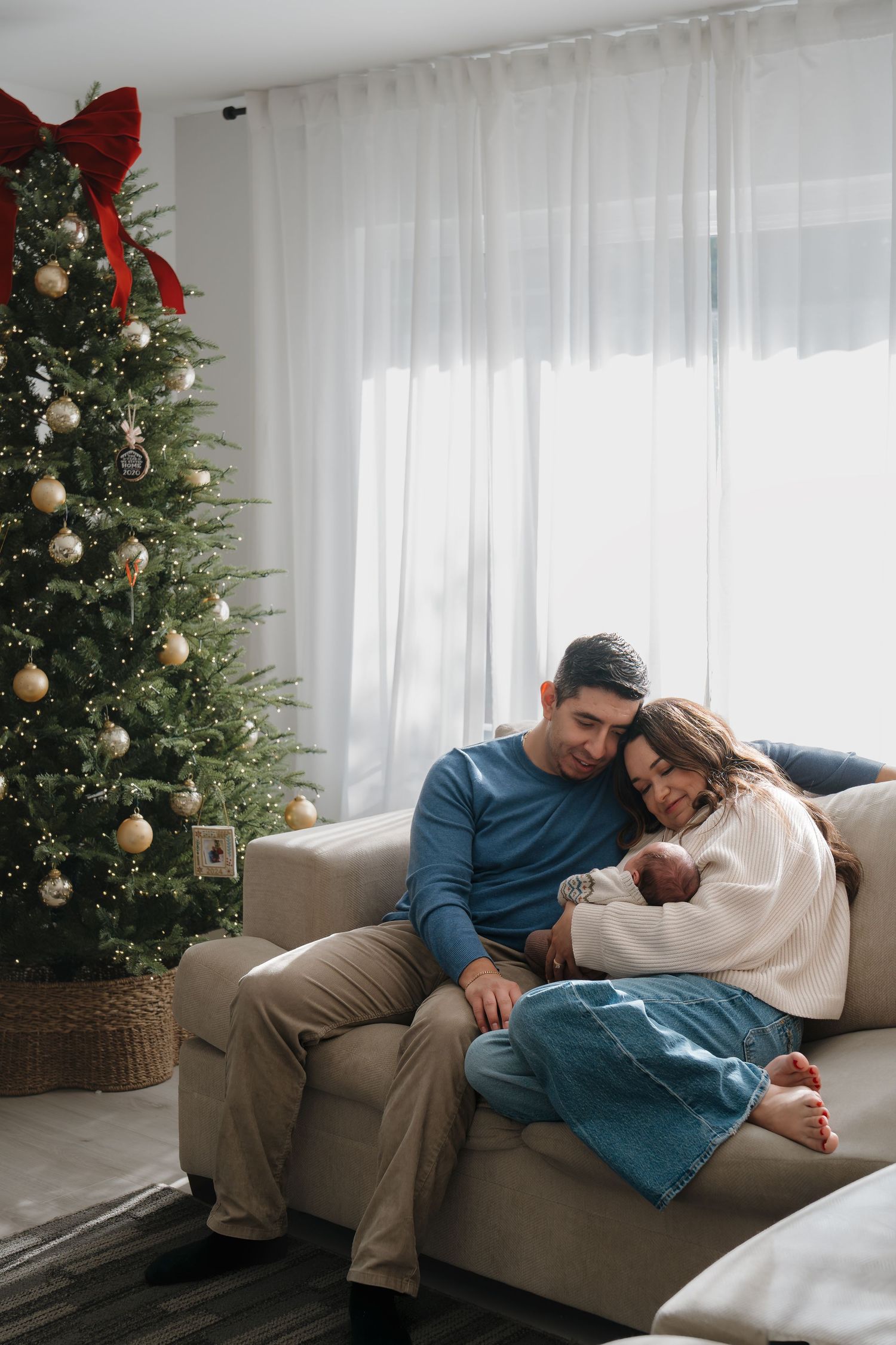Cozy Christmas scene showing couple relaxing on beige couch near decorated tree with gold ornaments and white curtains.