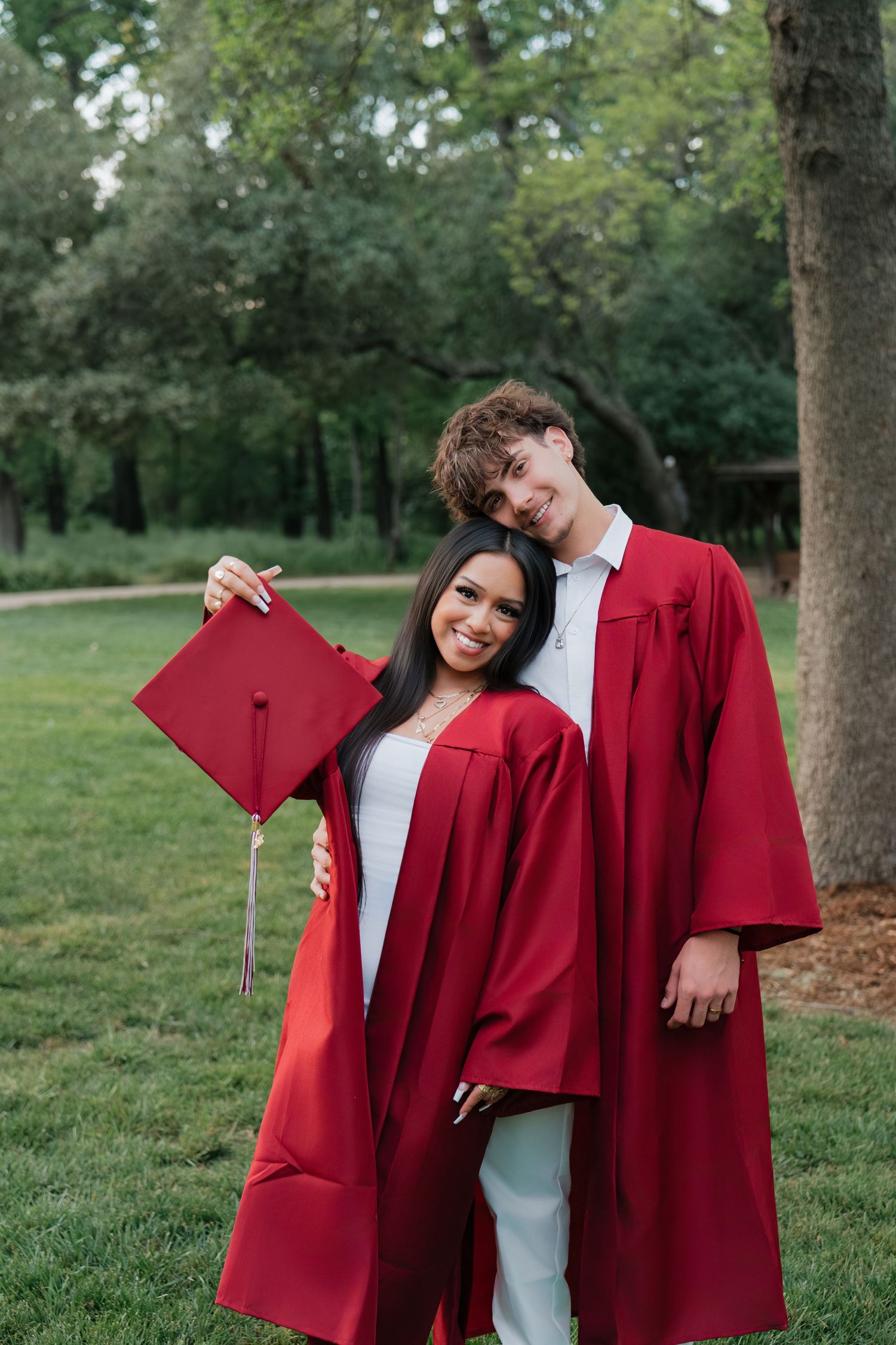 Two graduates in red caps and gowns smiling together outdoors in a lush green park setting.